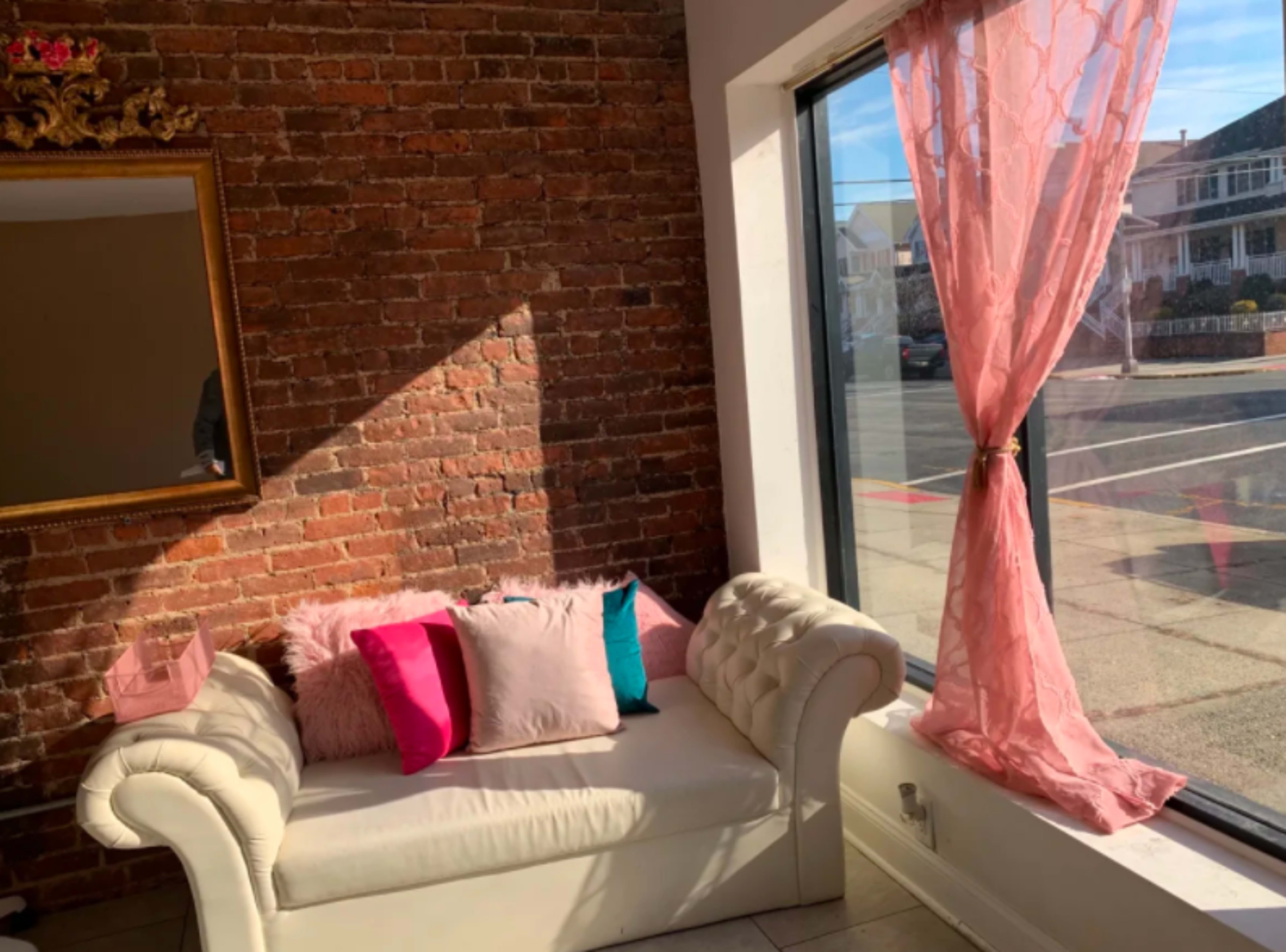 A white couch with colorful pillows is positioned near a large window with sheer pink curtains, beside a brick wall.