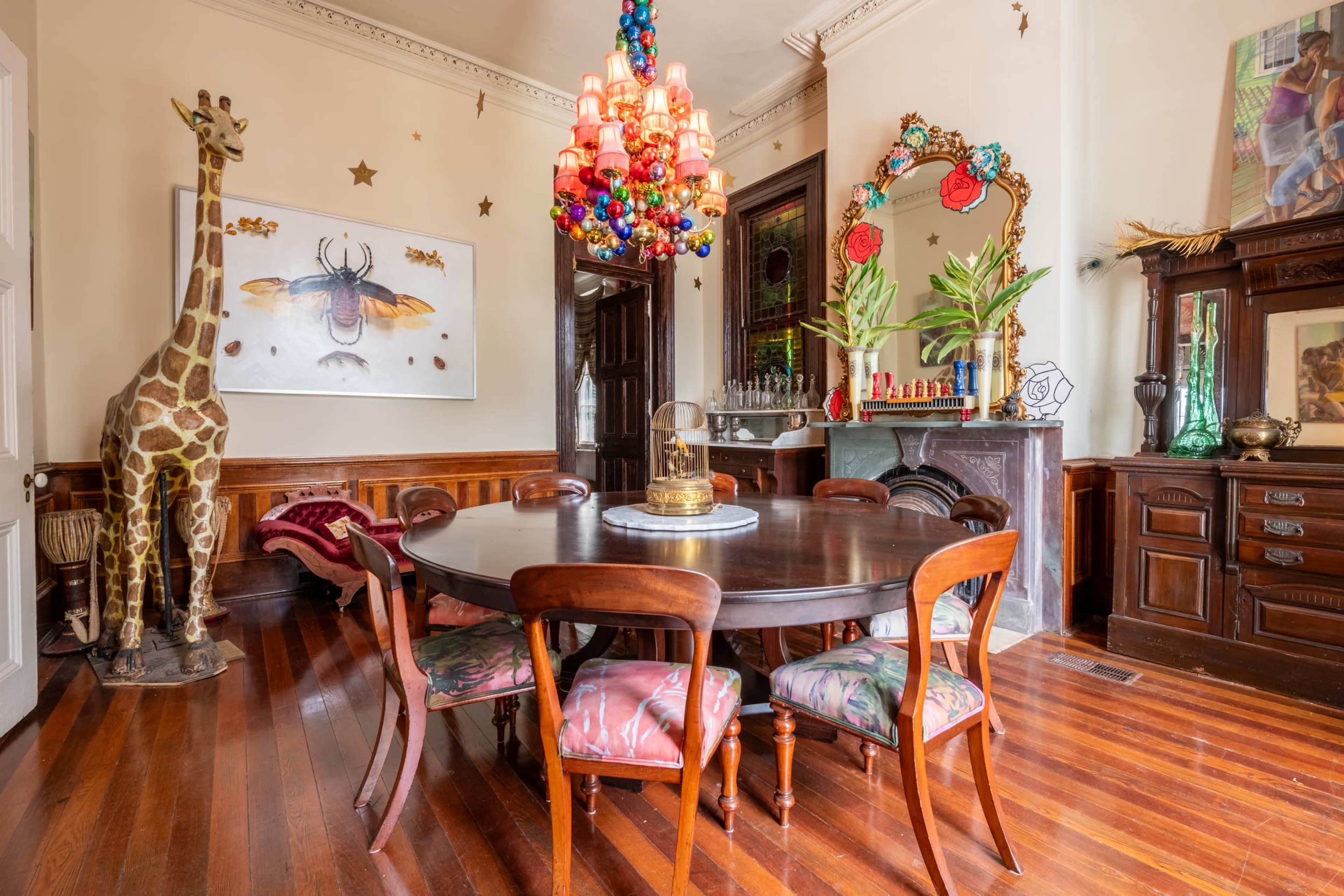 The room features a round dining table surrounded by chairs, with a tall giraffe sculpture in one corner and a colorful chandelier overhead.