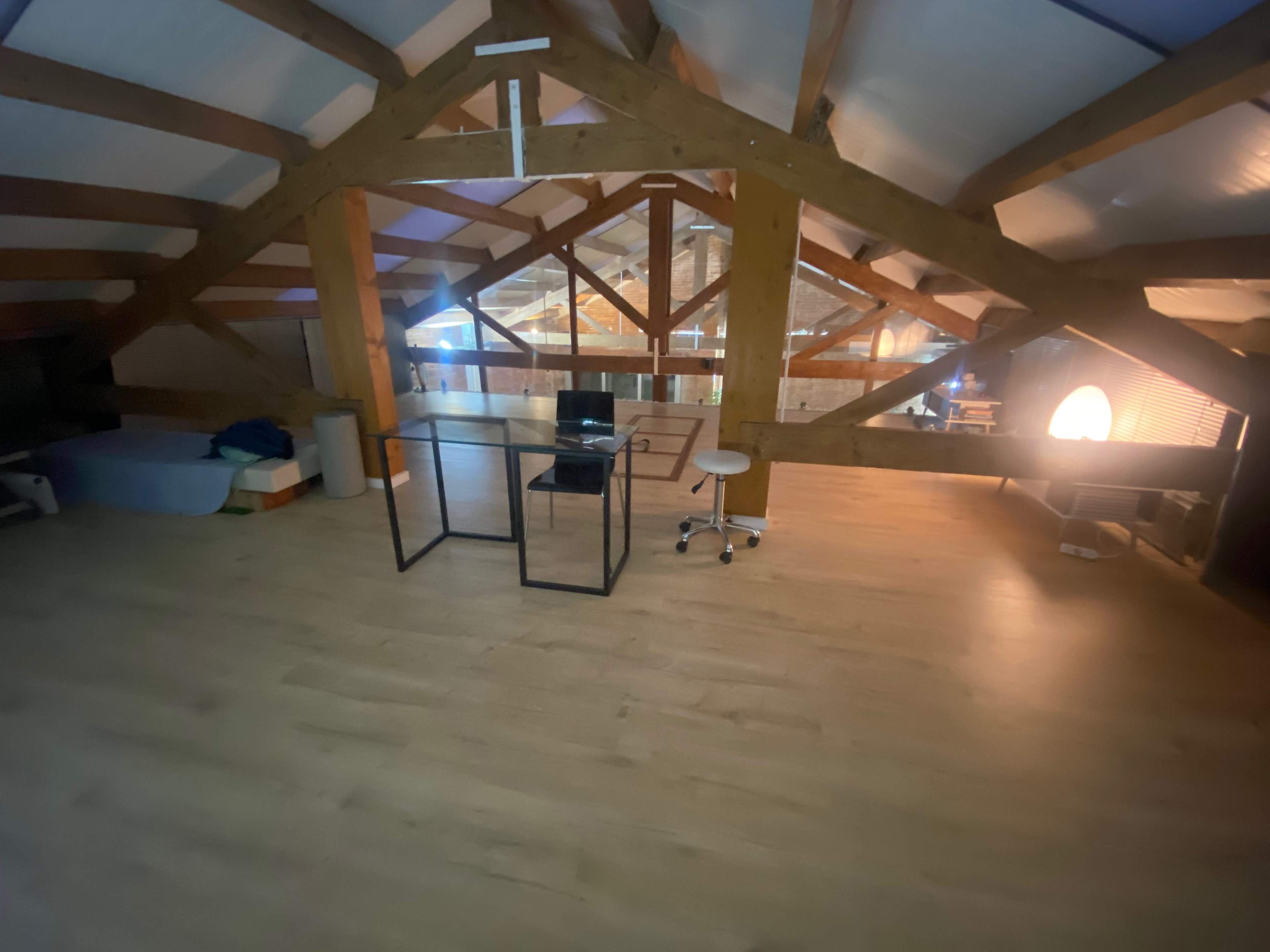 The image shows a spacious loft with wooden beams, a pale wooden floor, a computer setup on a glass table, and a few chairs.