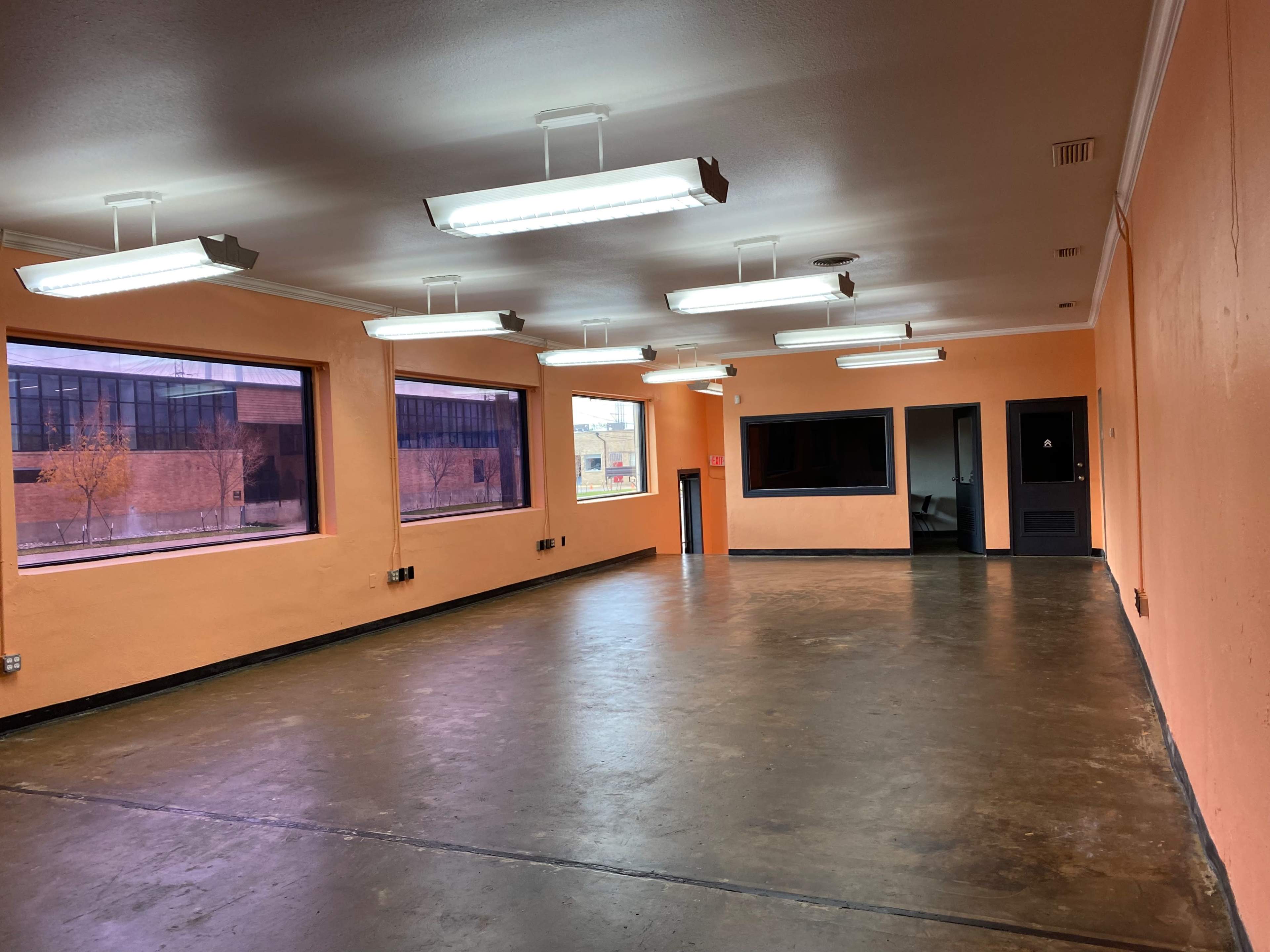 An empty room with orange walls, fluorescent lights, and large windows letting in natural light.