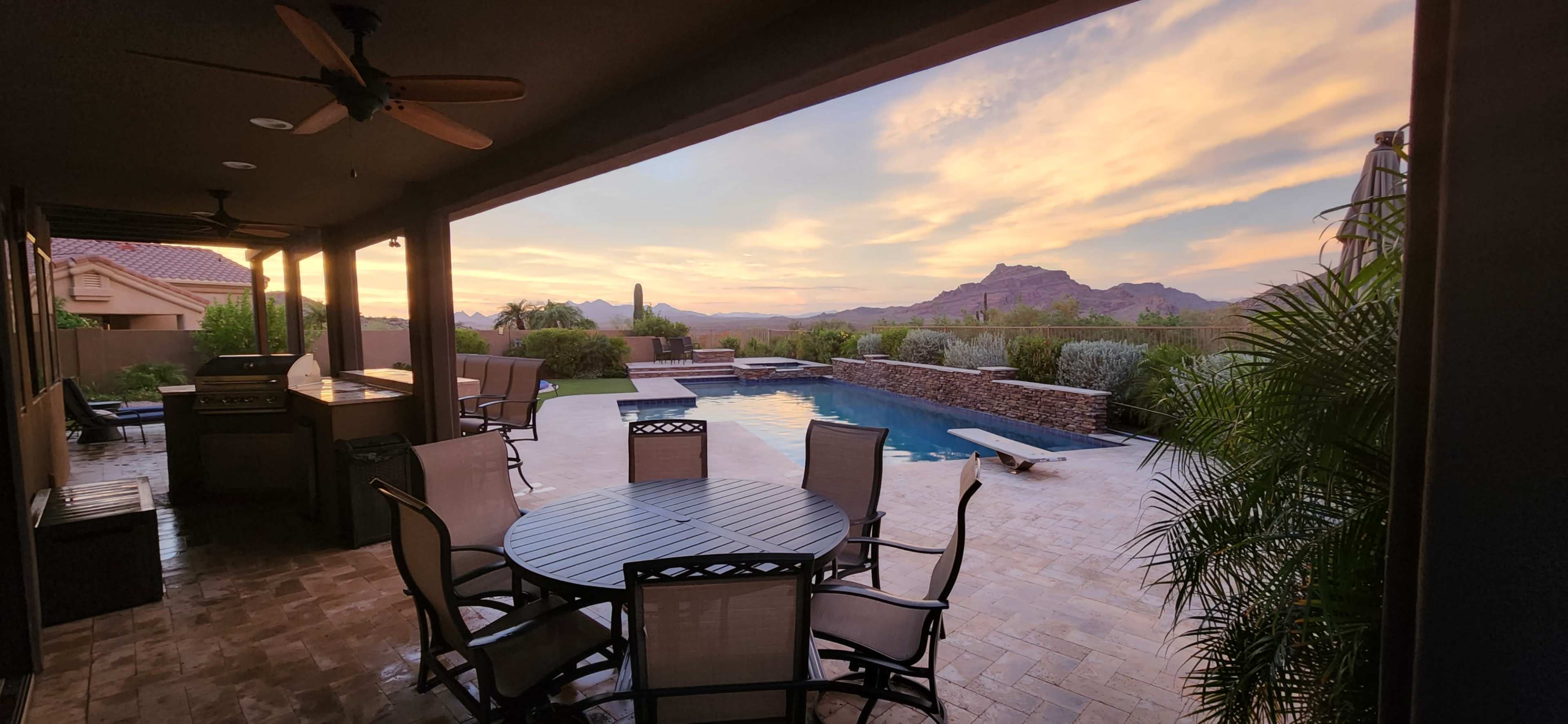 The image shows a covered patio with a dining table and chairs overlooking a swimming pool and mountains at sunset.