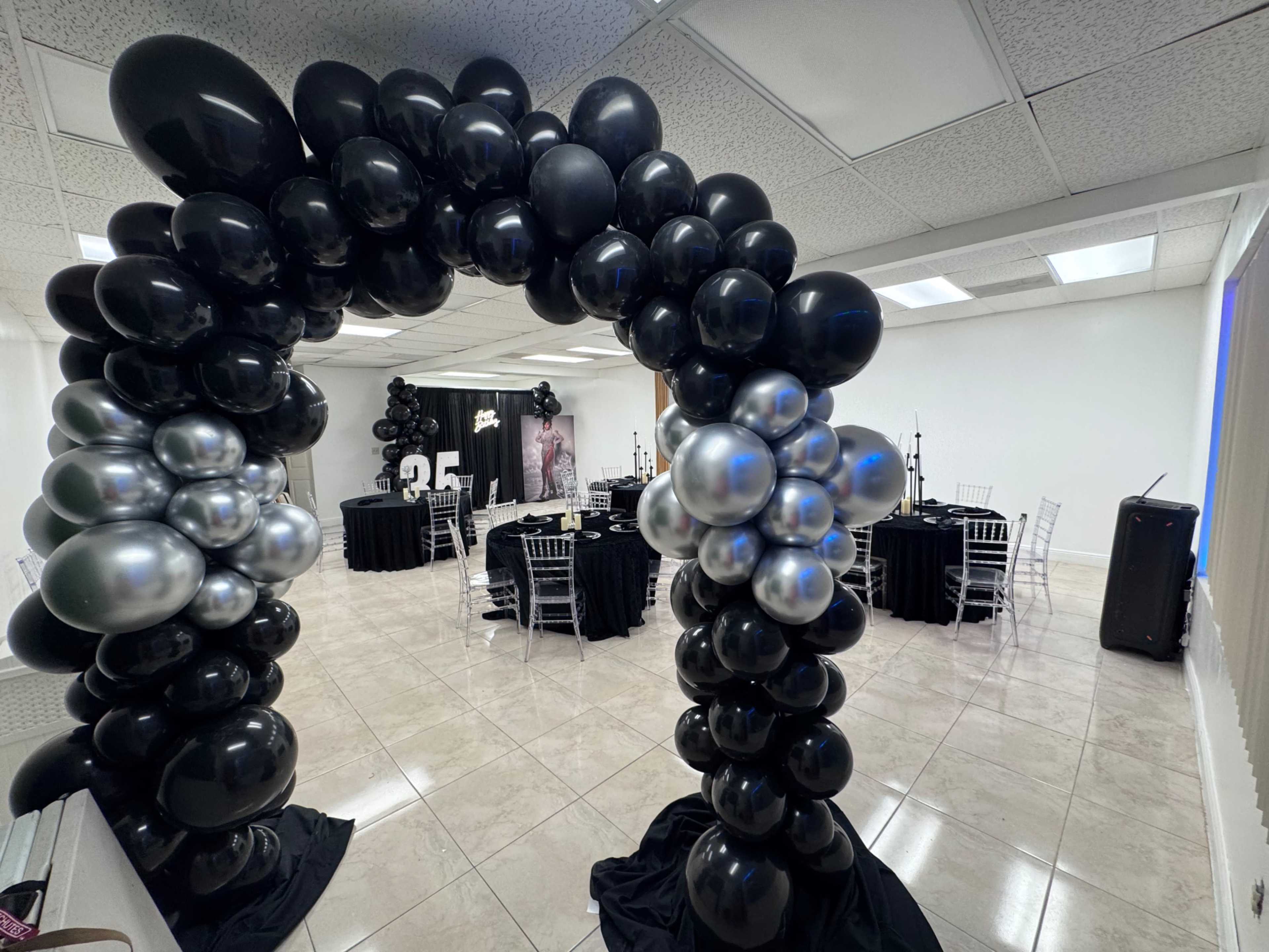 The image shows a decorated event space featuring an arch made of black and silver balloons, with black tablecloths and transparent chairs arranged for a celebration.