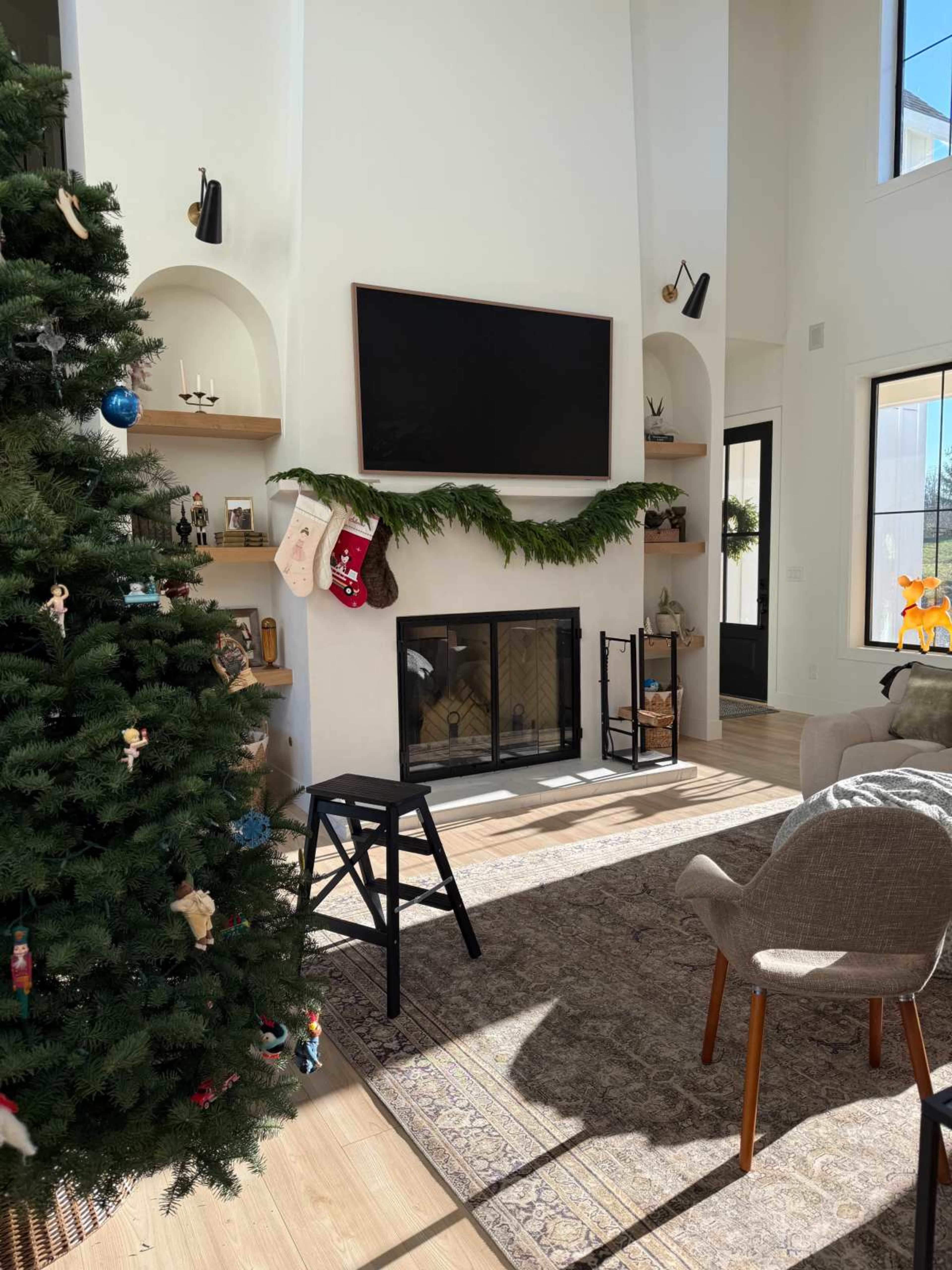 The image shows a bright living room decorated for the holidays, featuring a Christmas tree on the left, a fireplace with stockings hanging, and a television mounted on the wall.