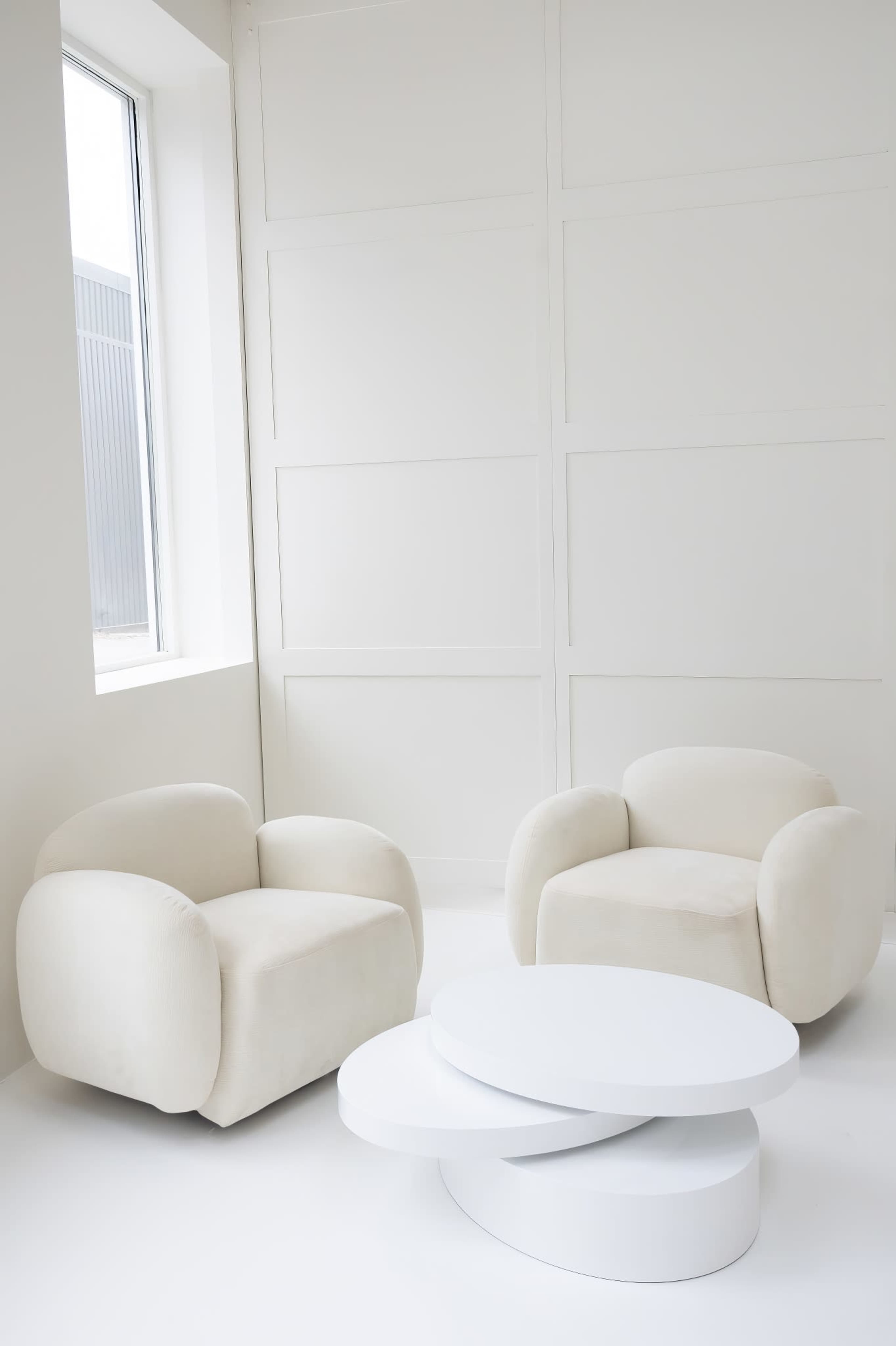 Two cream-colored, curved armchairs facing a white, modern coffee table in a well-lit, minimalistic room.