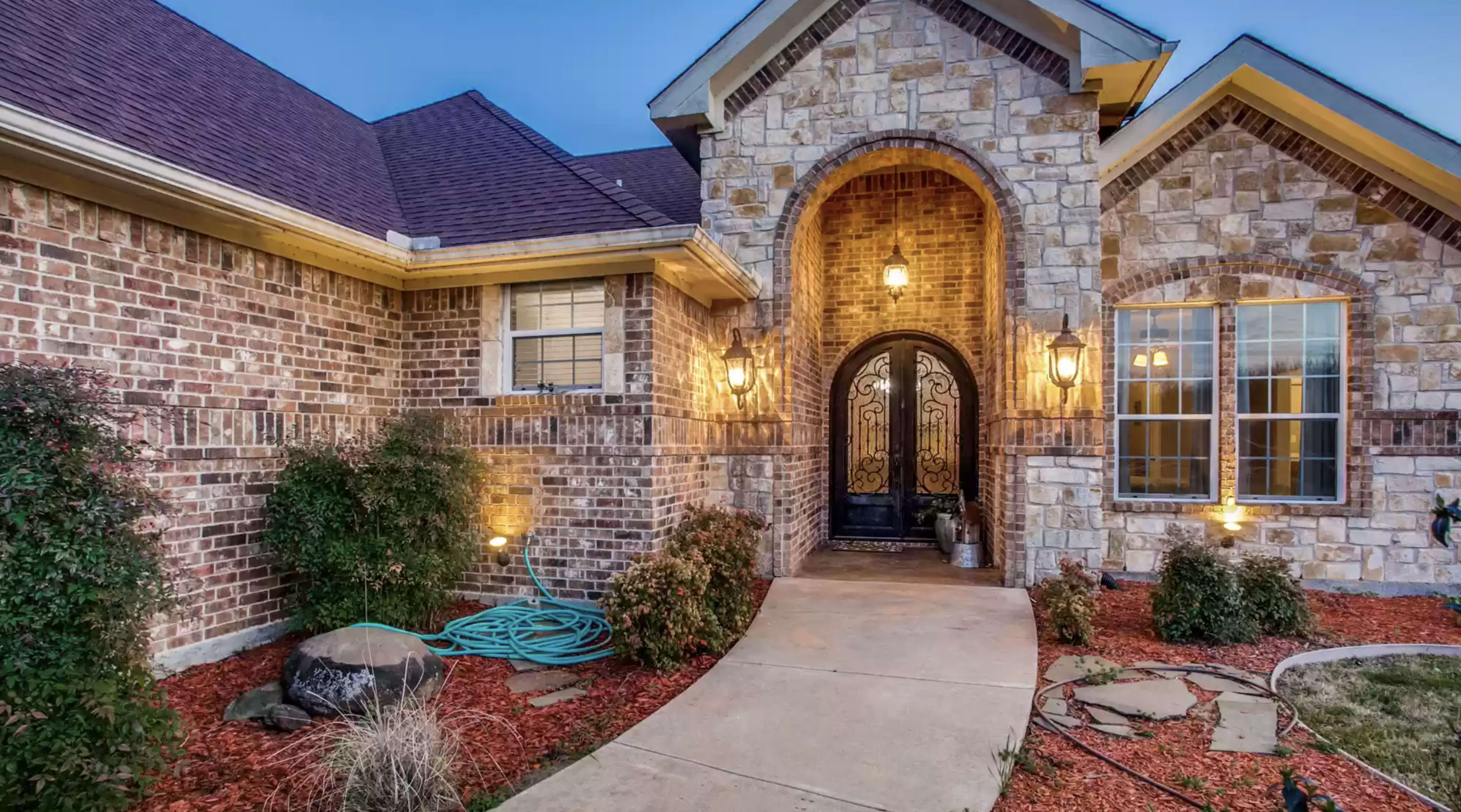 A brick and stone single-story home with a curved pathway leading to a decorative front door, framed by landscaped flower beds and outdoor lighting.