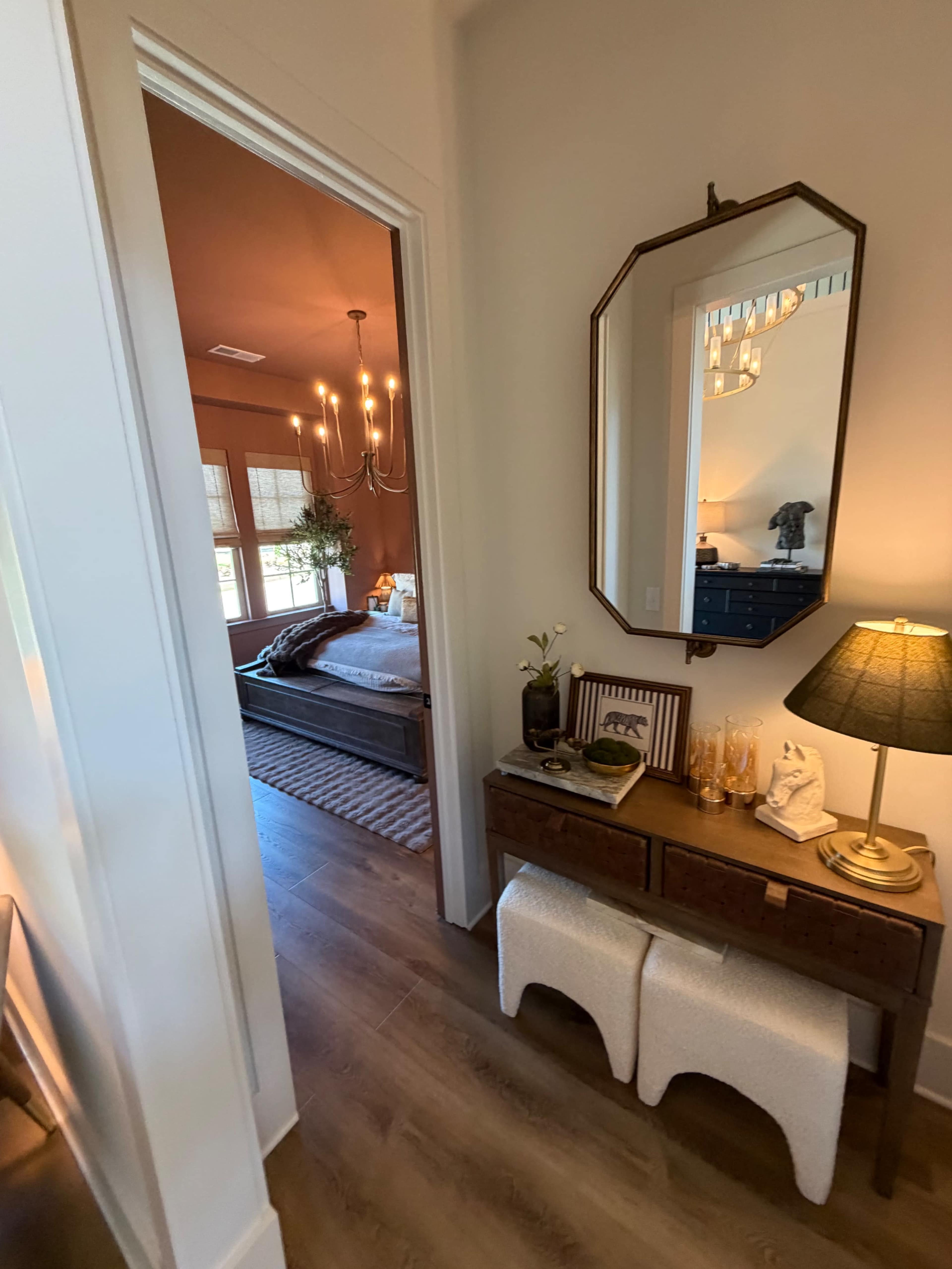 The image shows a hallway leading to a well-decorated bedroom with a chandelier, a bed, and a dark-toned piece of furniture, while a small console table with a lamp and decorative items is positioned in the foreground.