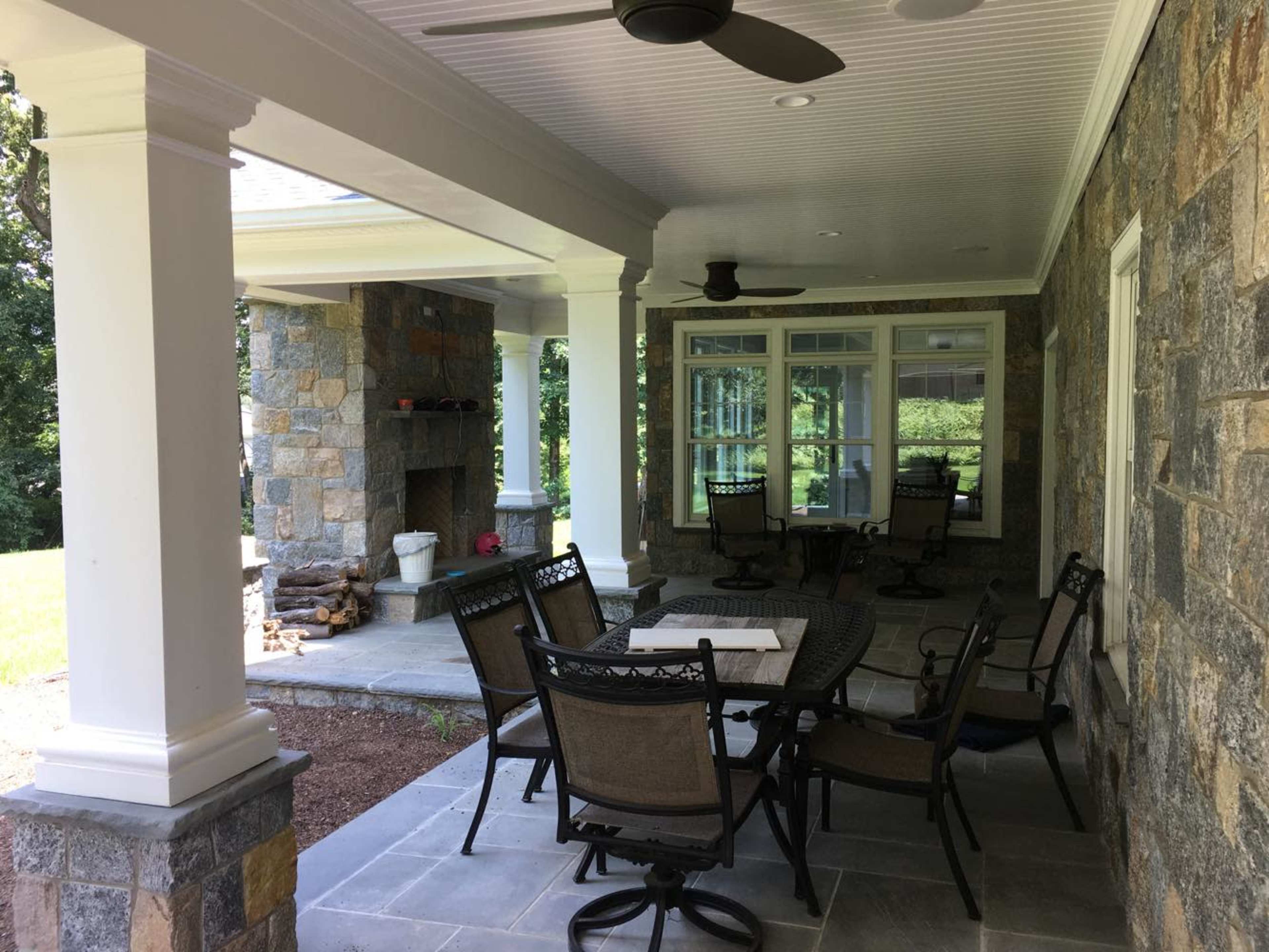 The image shows a covered outdoor patio with a stone fireplace, table, and seating surrounded by lawn and trees.