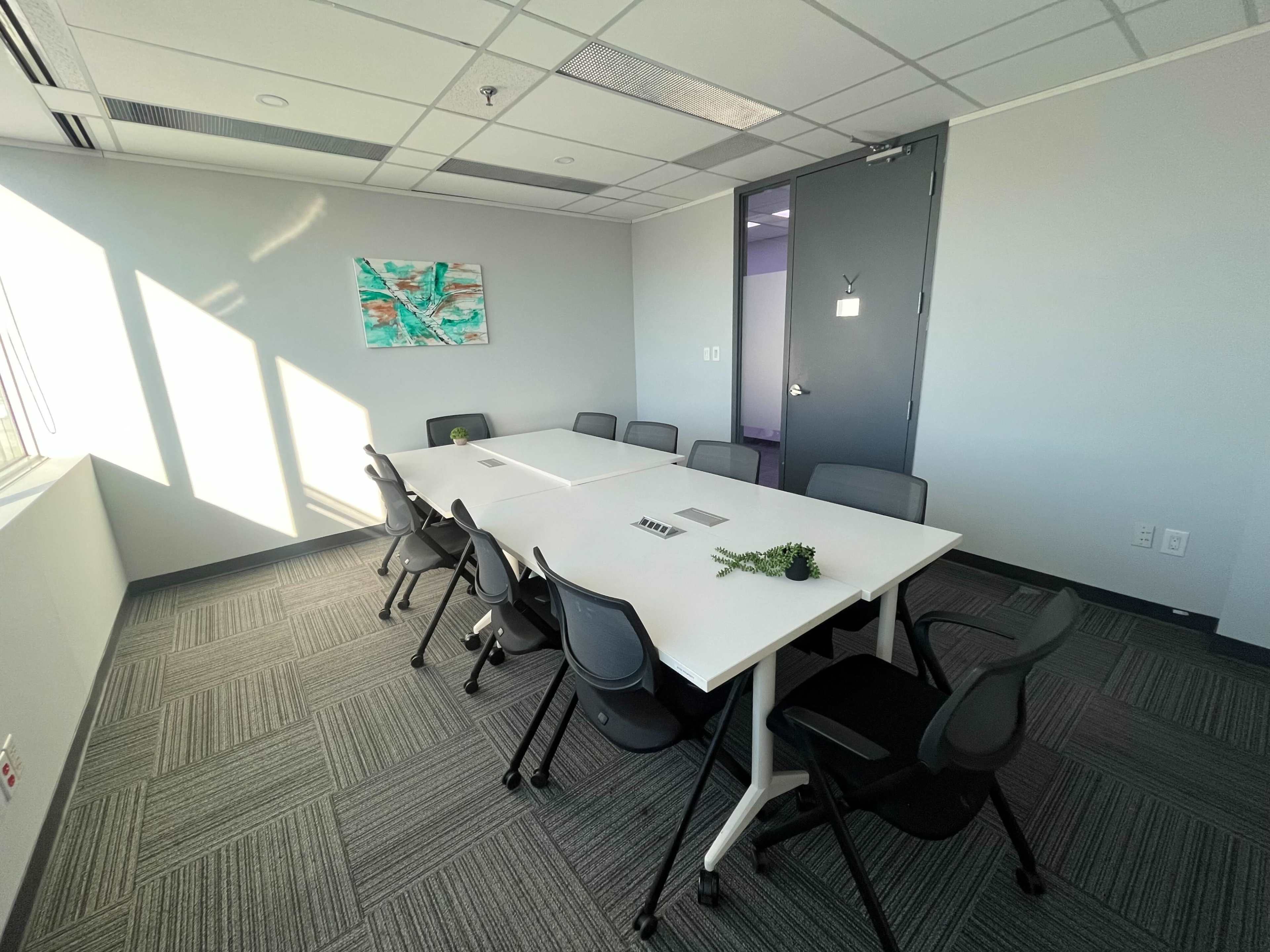 A modern conference room features a large white table surrounded by black chairs, with a wall art piece and natural light coming through a window.