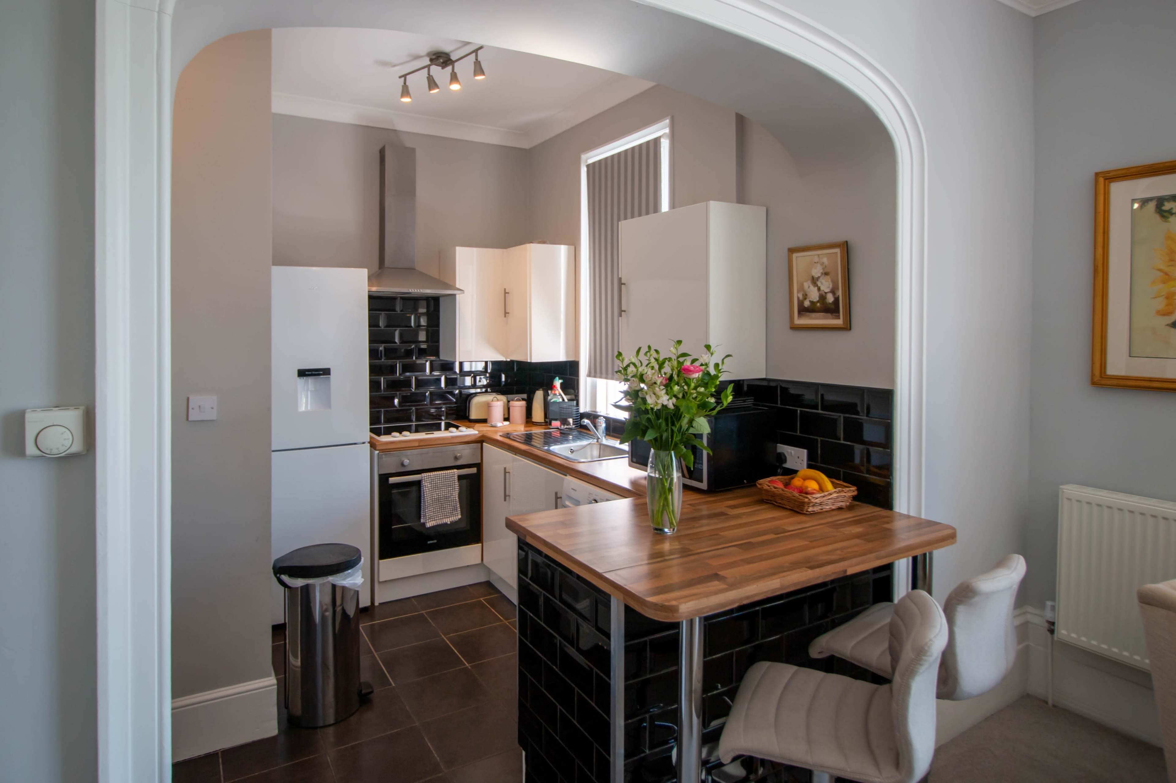 A modern kitchen features white cabinets, a black tiled backsplash, and a wooden table with a vase of flowers and a bowl of fruit.