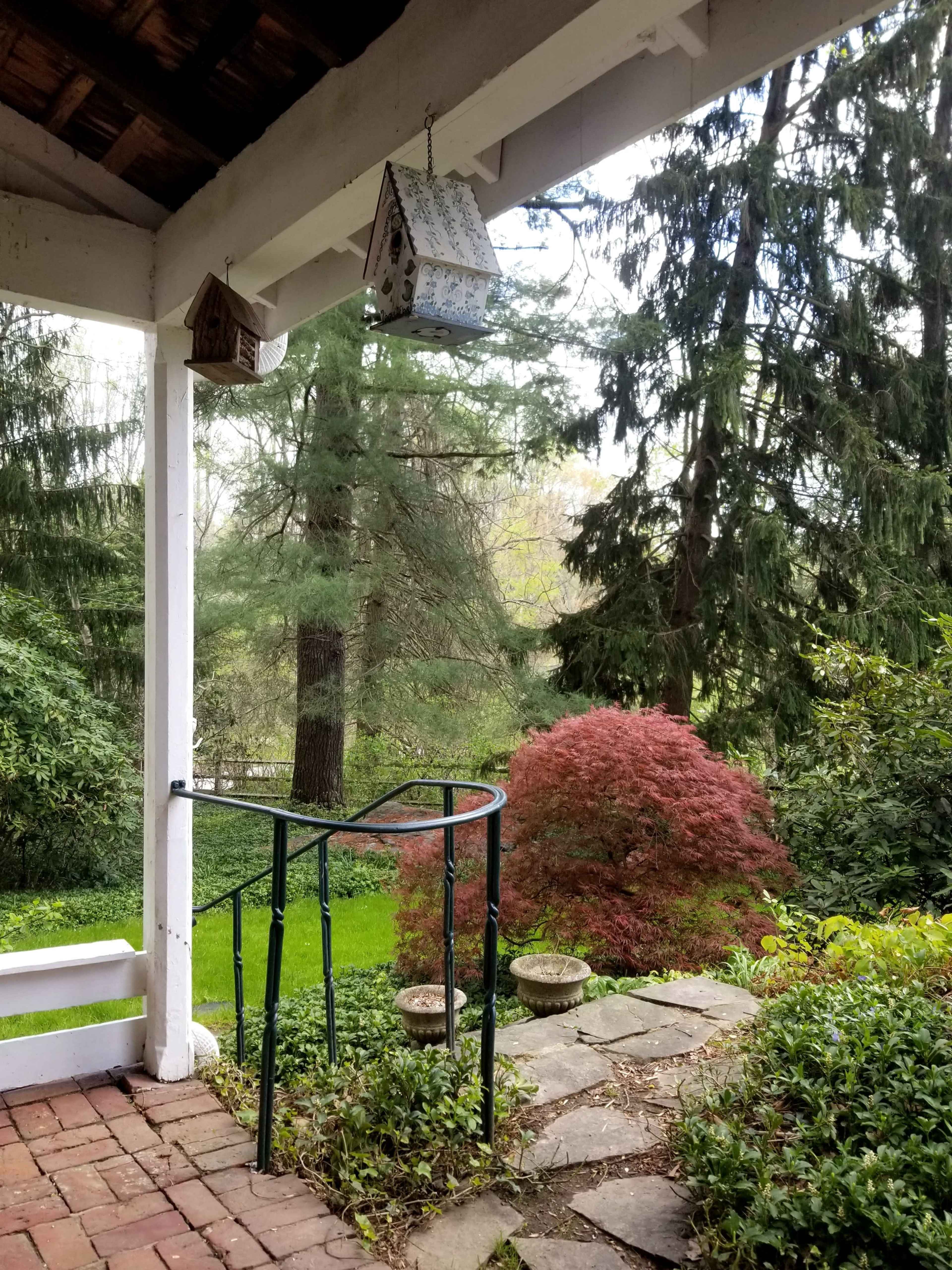A view from a covered porch, featuring a curved metal railing, a stone pathway, and various trees and shrubs in the garden.