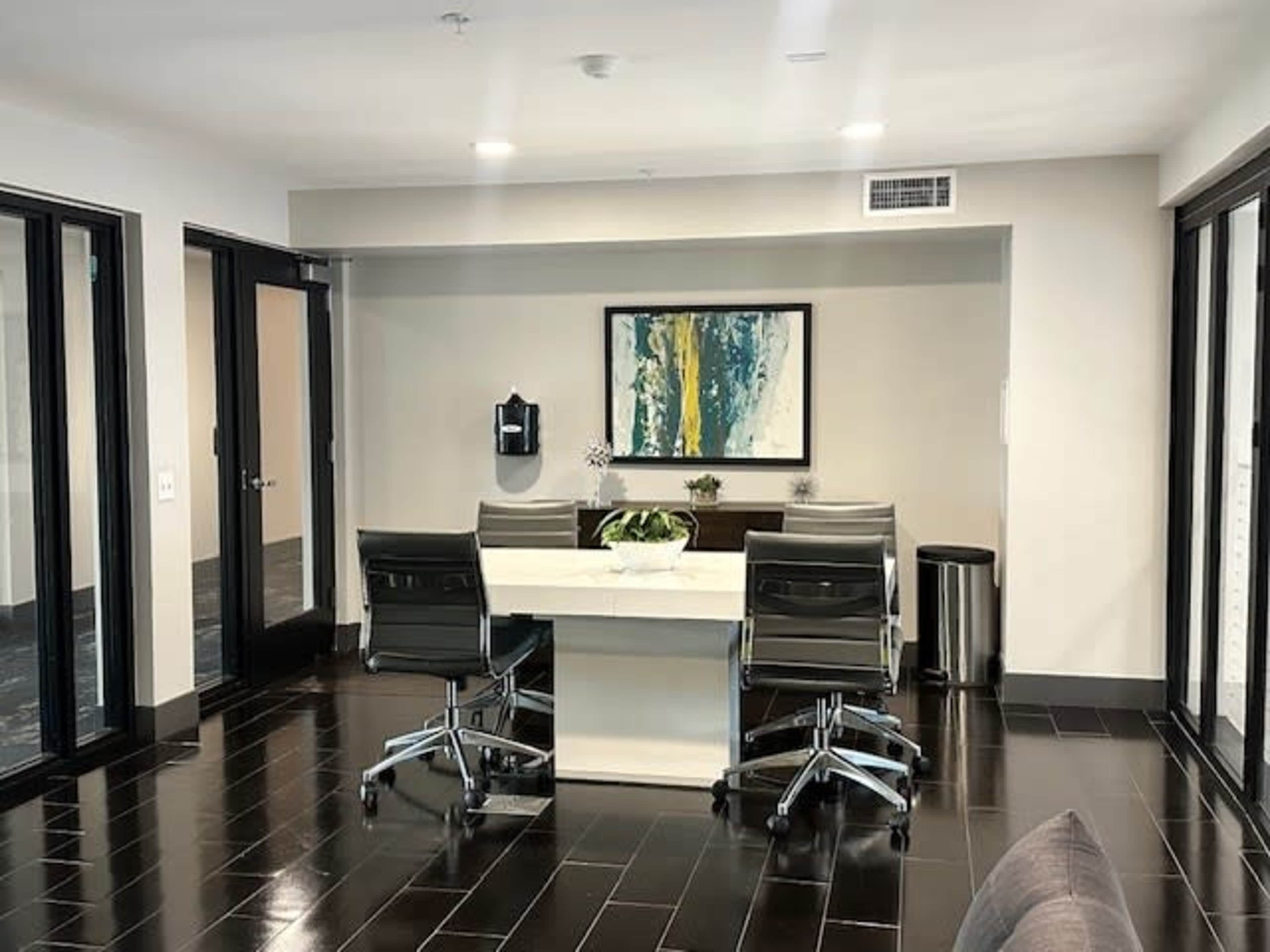 The image shows a modern office space featuring a rectangular table with four black chairs, a wall-mounted dispenser, a potted plant centerpiece, and large windows.