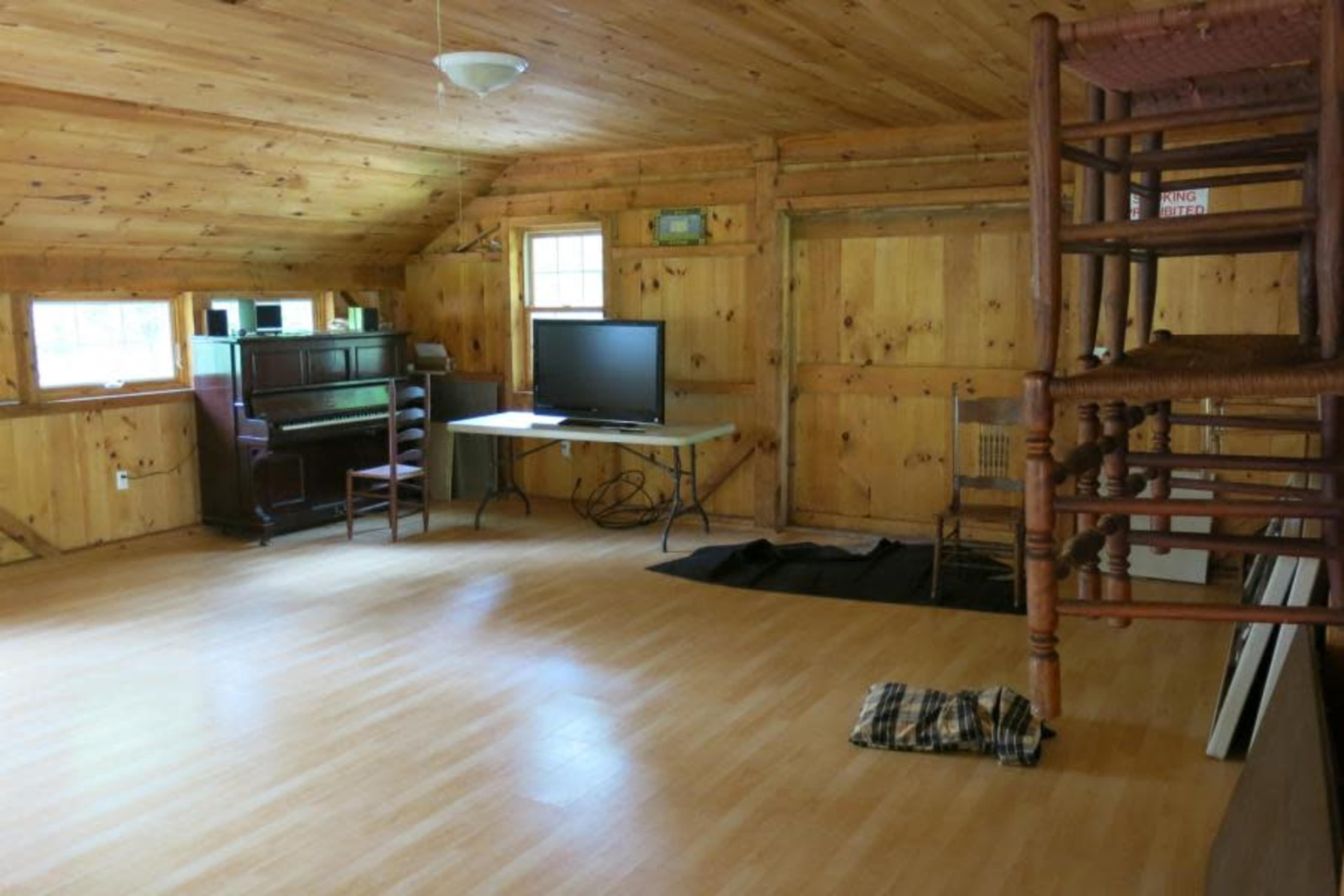 The interior of a wooden room features a piano, a television on a table, and stacked chairs against the wall.