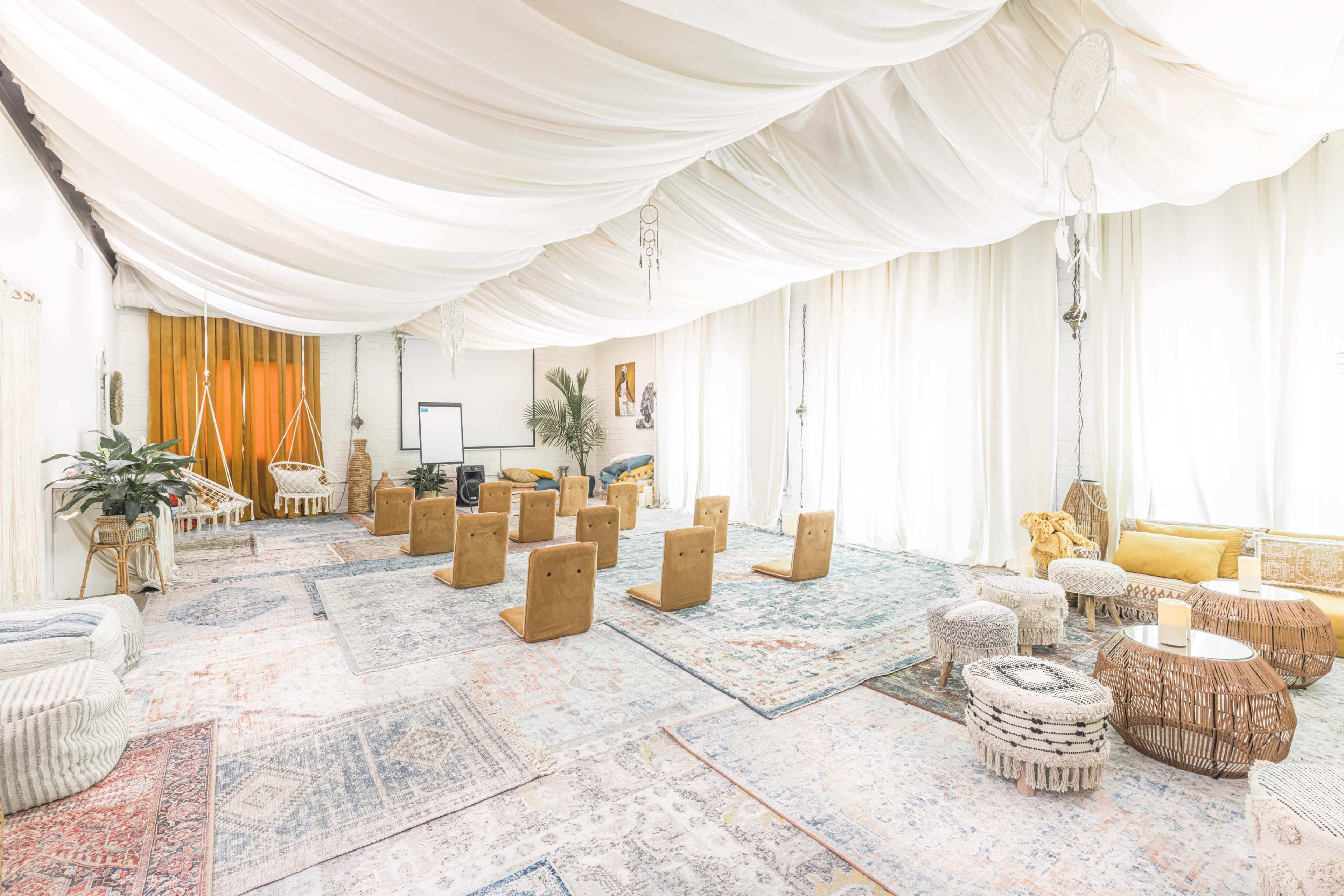 A spacious, well-lit room is arranged with wooden seating and various decorative elements on colorful rugs beneath a draped ceiling.