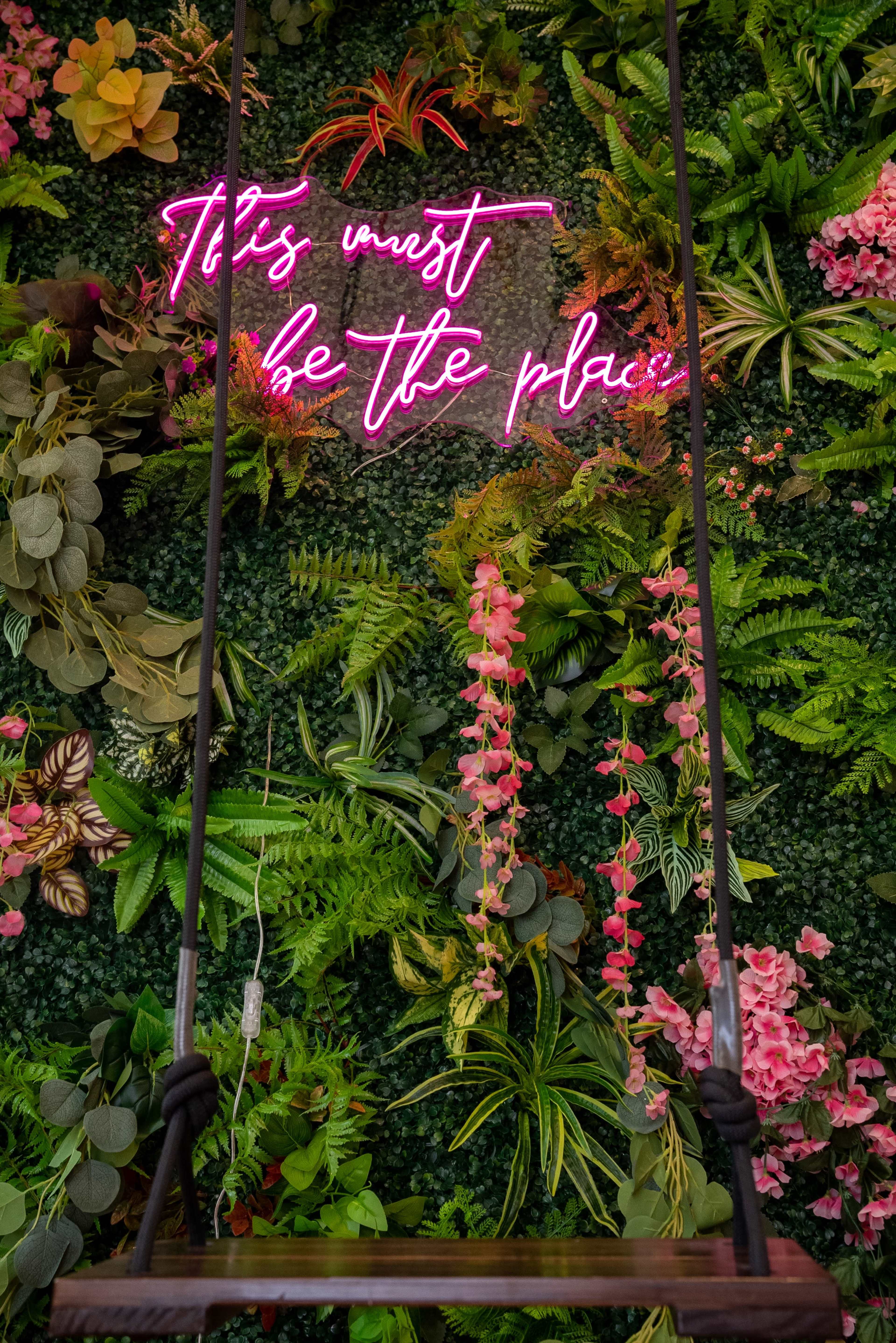 A swing hangs in front of a vibrant green wall adorned with various plants and a neon sign that reads, "It must be the place."