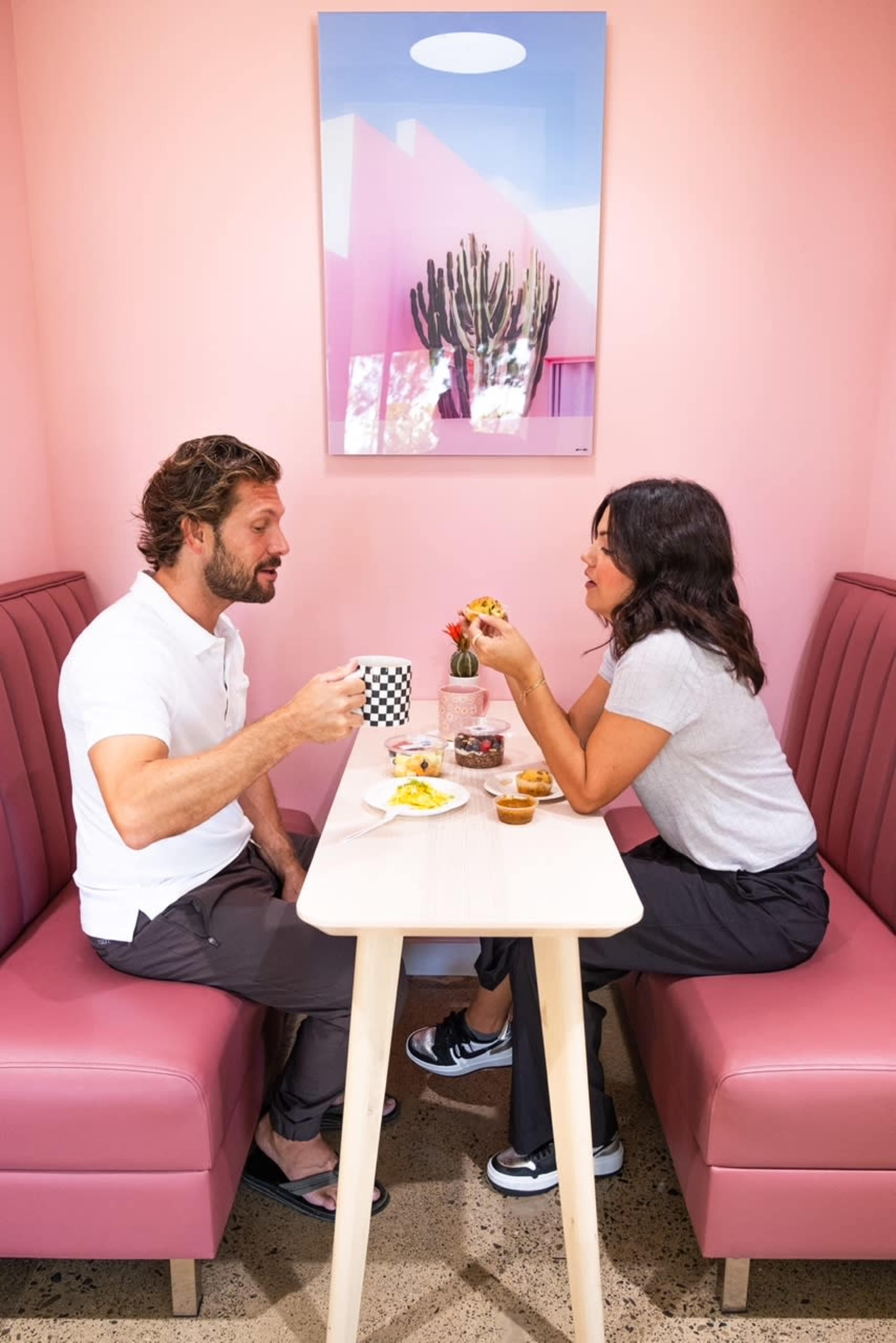 A man and a woman sit at a pink booth in a café, enjoying food and drinks while engaged in conversation.