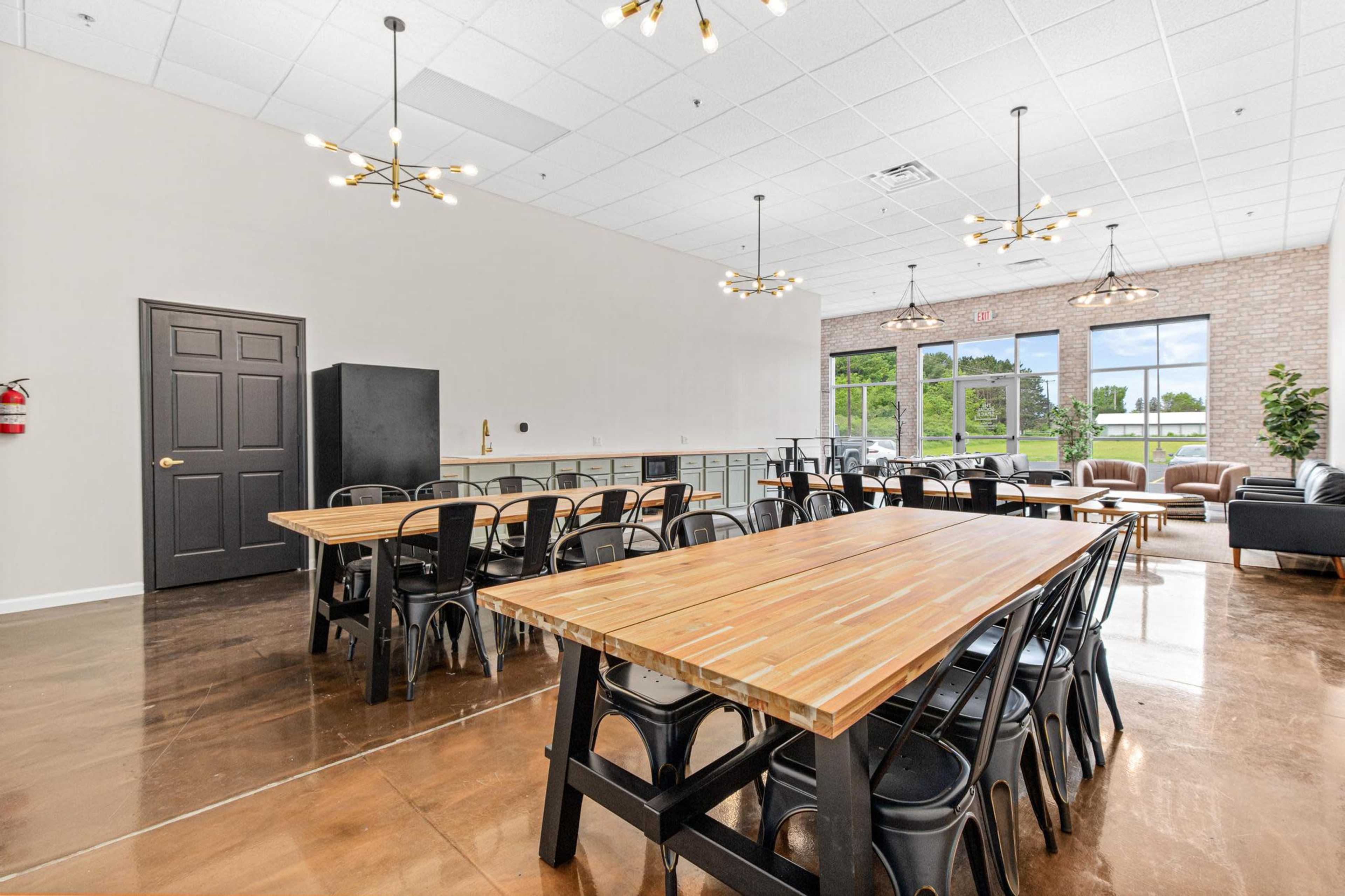A spacious dining area with long wooden tables and metal chairs, featuring large windows that provide natural light and a view of greenery outside.