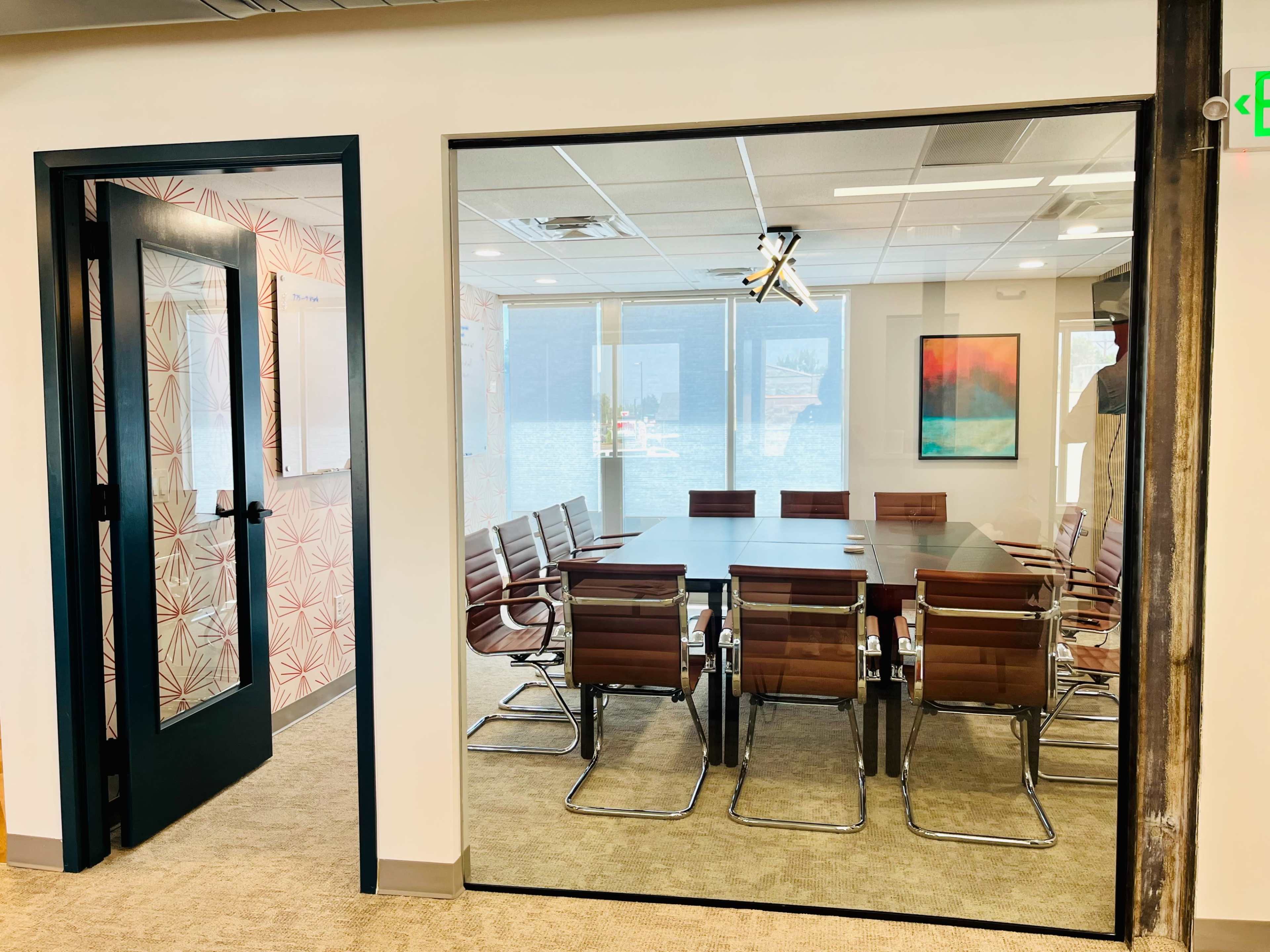 A modern conference room with a long table and several chairs, viewed through a glass wall from an adjacent hallway.