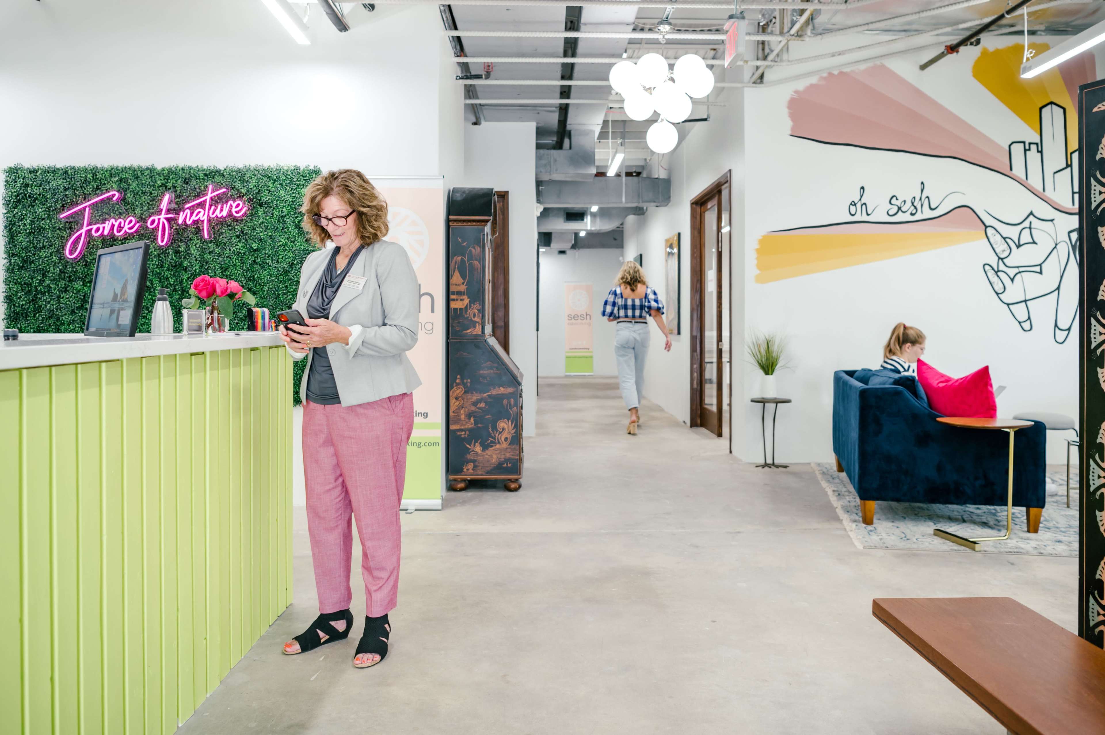 A woman in business attire stands at a reception desk with a vibrant green and pink theme, while another woman walks down the hallway and a third sits on a couch.