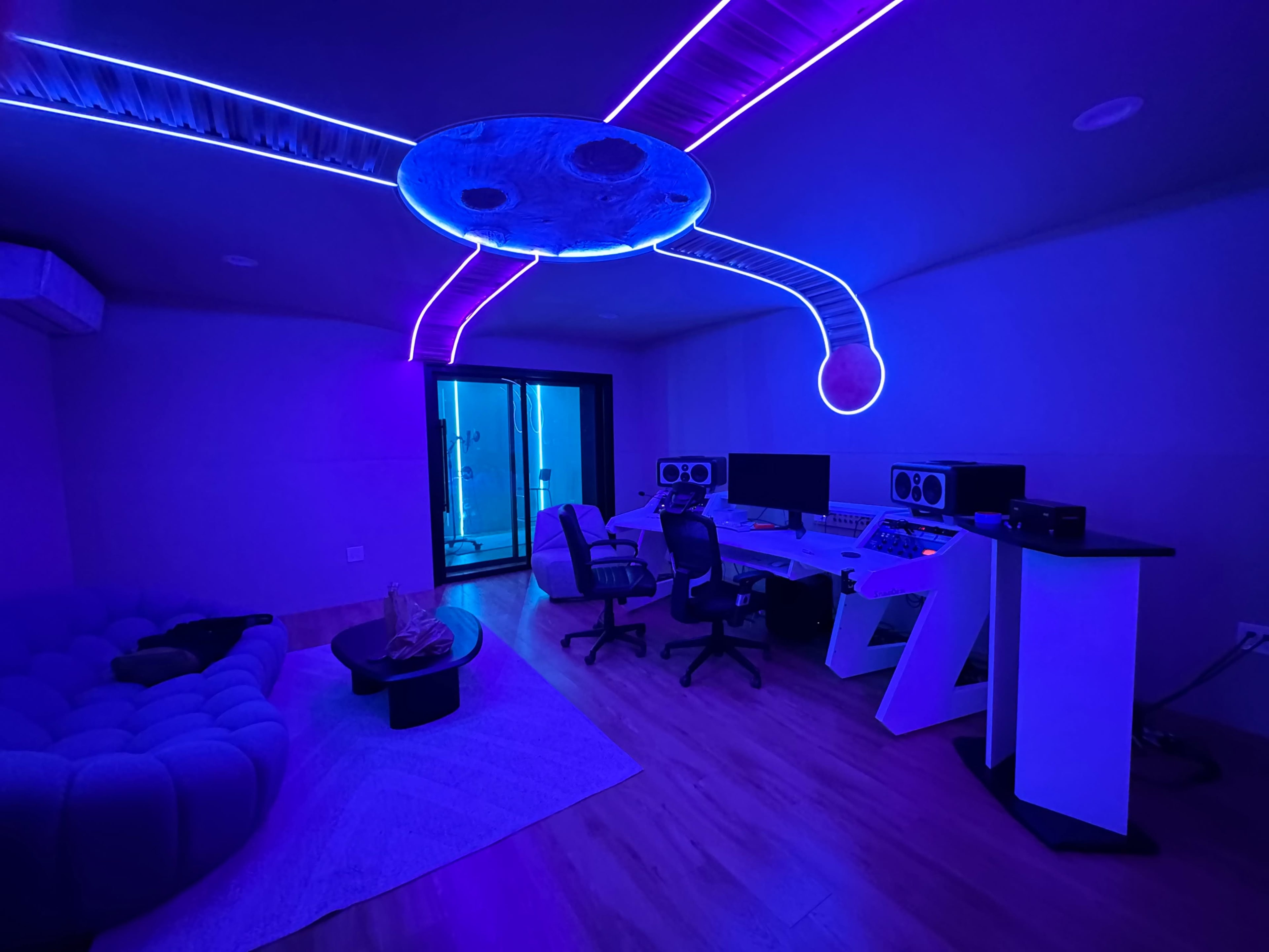 A modern music studio with purple LED lighting, featuring a large desk with audio equipment, two office chairs, and a cozy seating area.