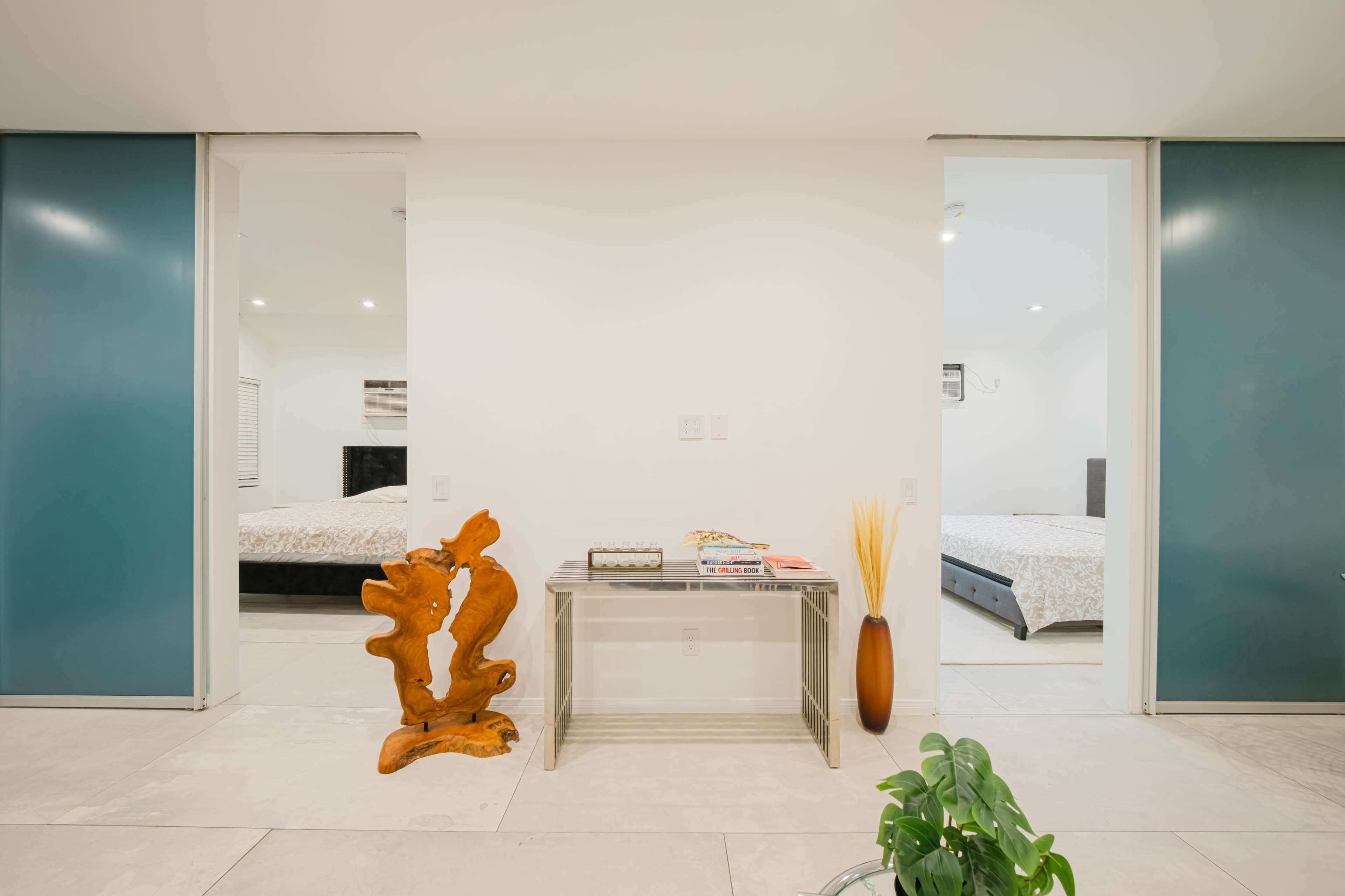 The image shows a modern, minimalist hallway featuring a wooden sculpture, a glass table, and two bedroom doors with beds partially visible through the open doorways.