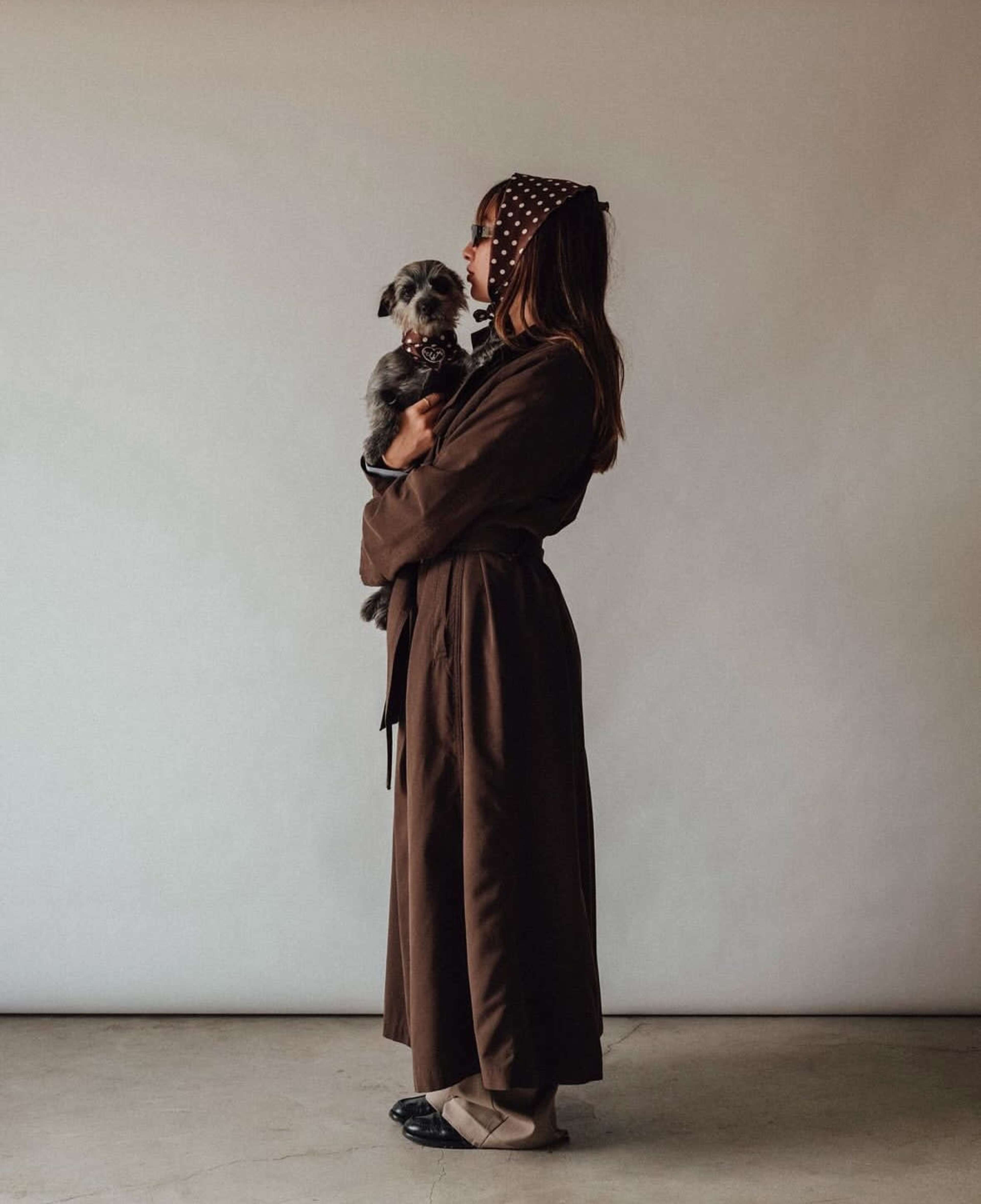 A woman in a brown robe holds a small dog while facing a plain light-colored background.