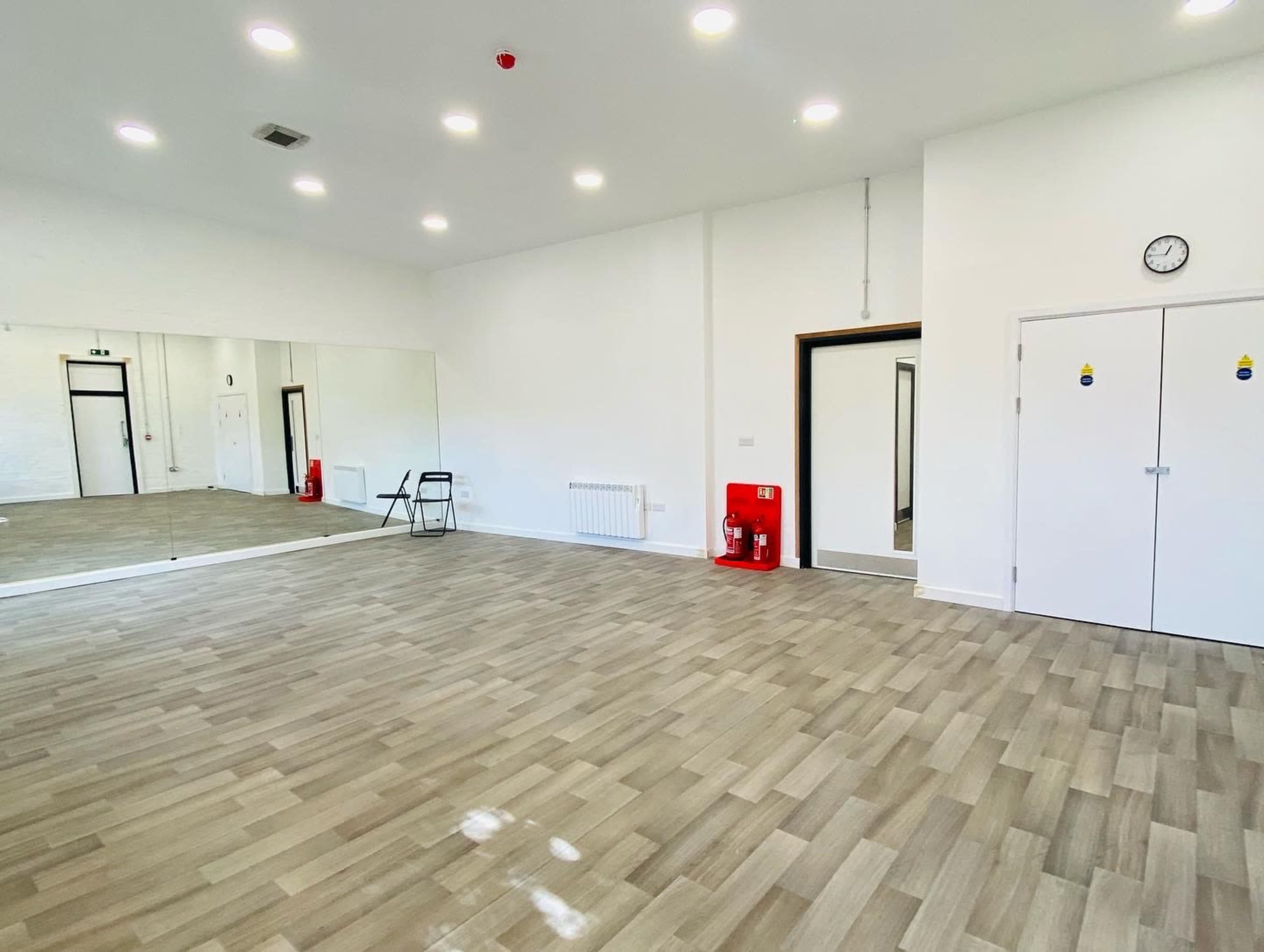 A spacious room with wooden flooring, a wall mirror, and a door leading to another area, along with a clock and fire safety equipment on the wall.