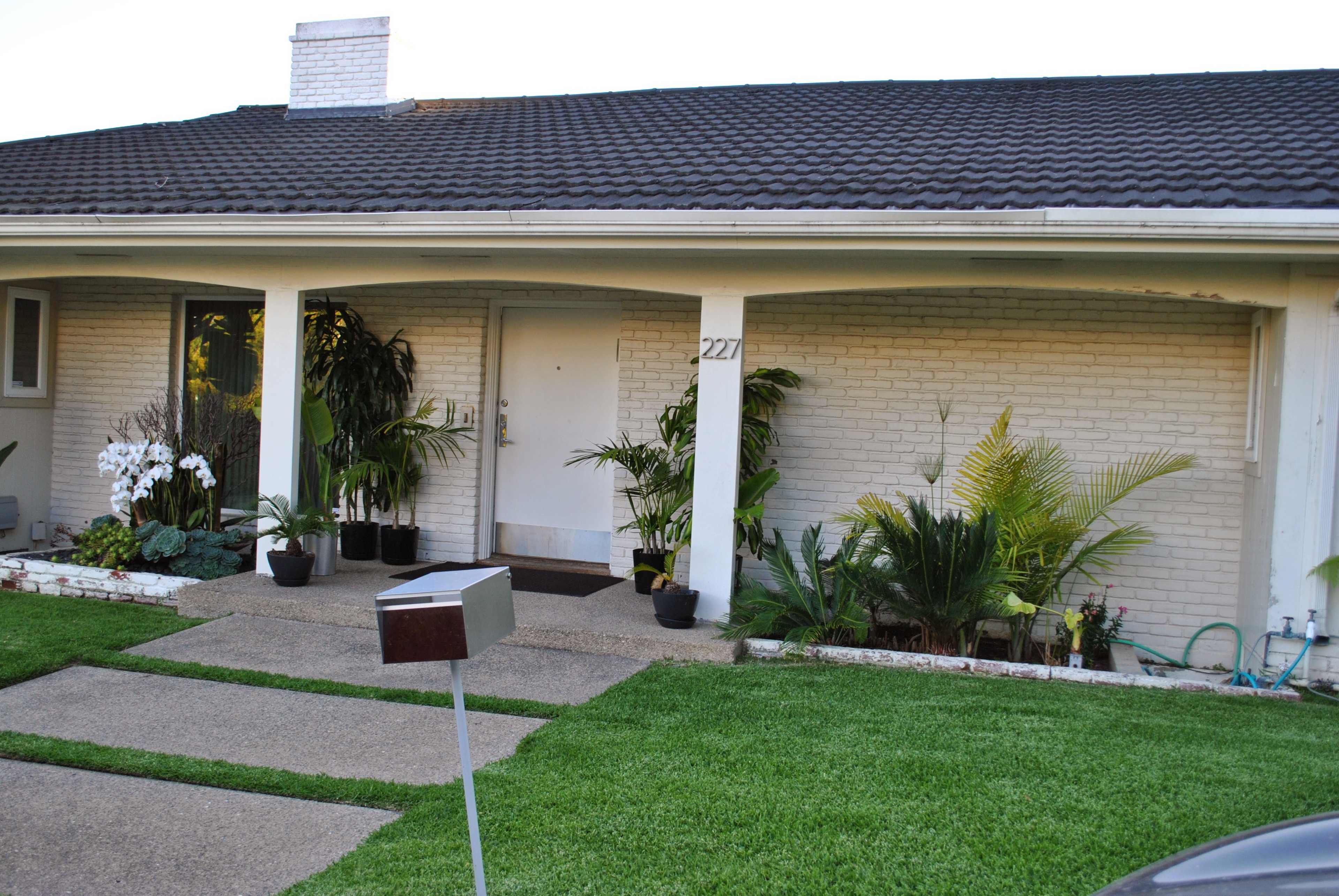A single-story house with a sloped roof features a front porch, a white door numbered 227, and potted plants lining the entrance.