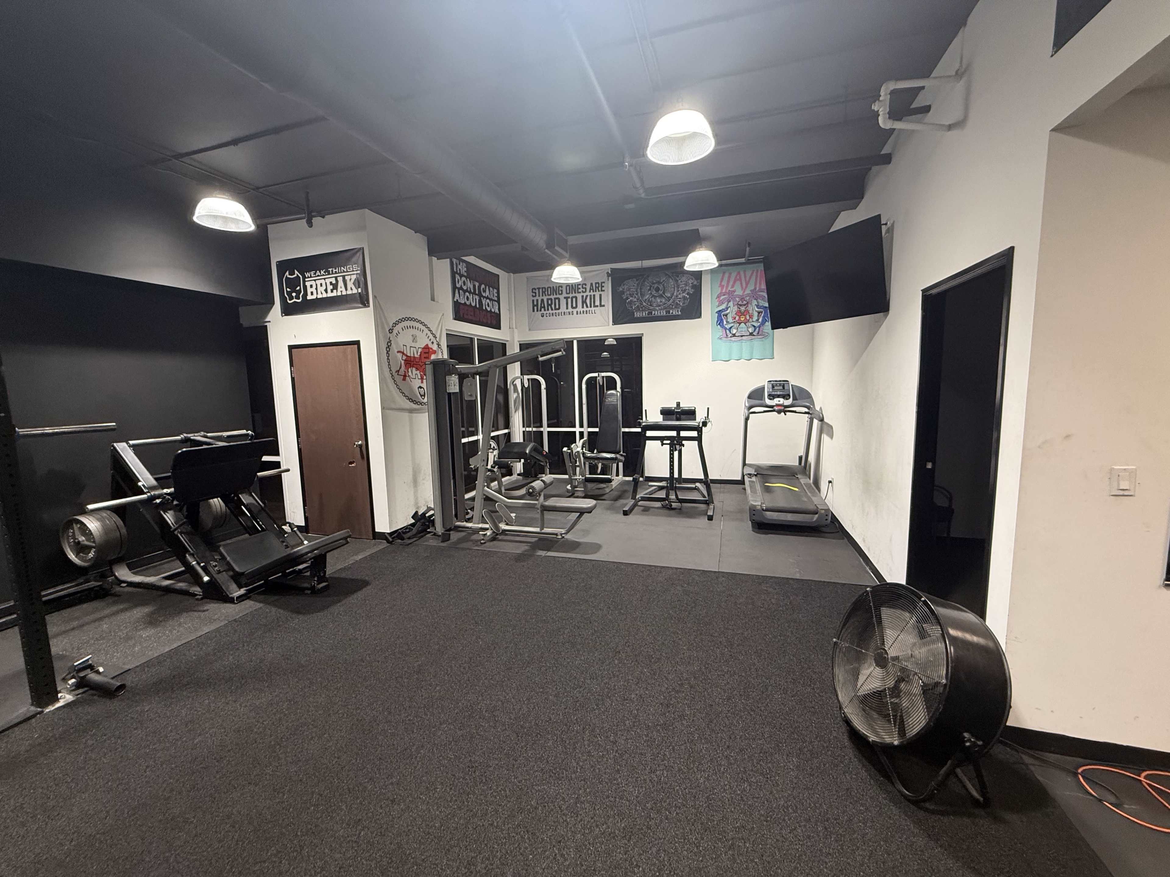 The image shows a gym interior with various fitness equipment, including weight machines, a treadmill, and a fan, on a black carpeted floor.