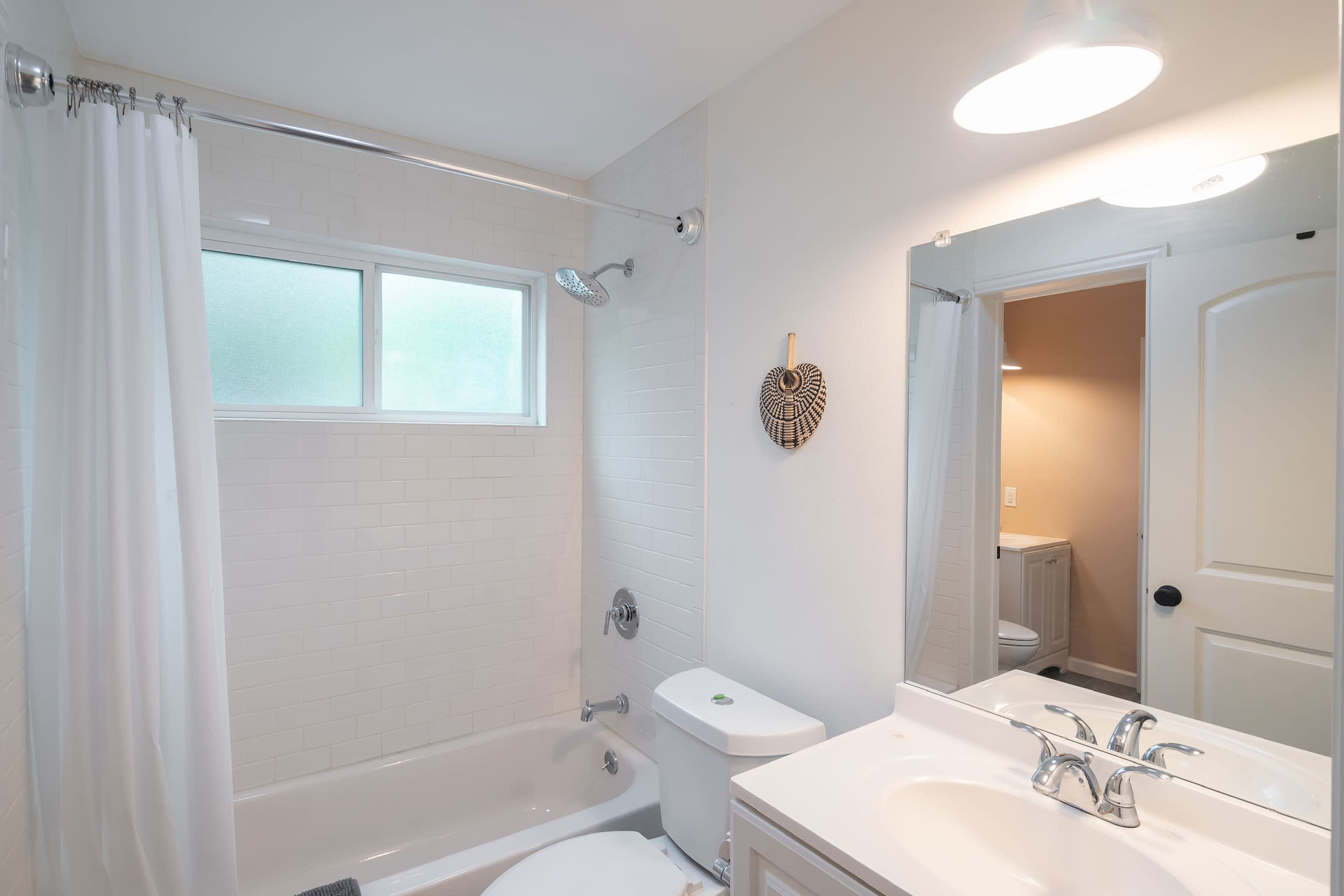The image shows a bathroom with a bathtub, shower, toilet, and a sink with a large mirror, featuring white tiles and a window.