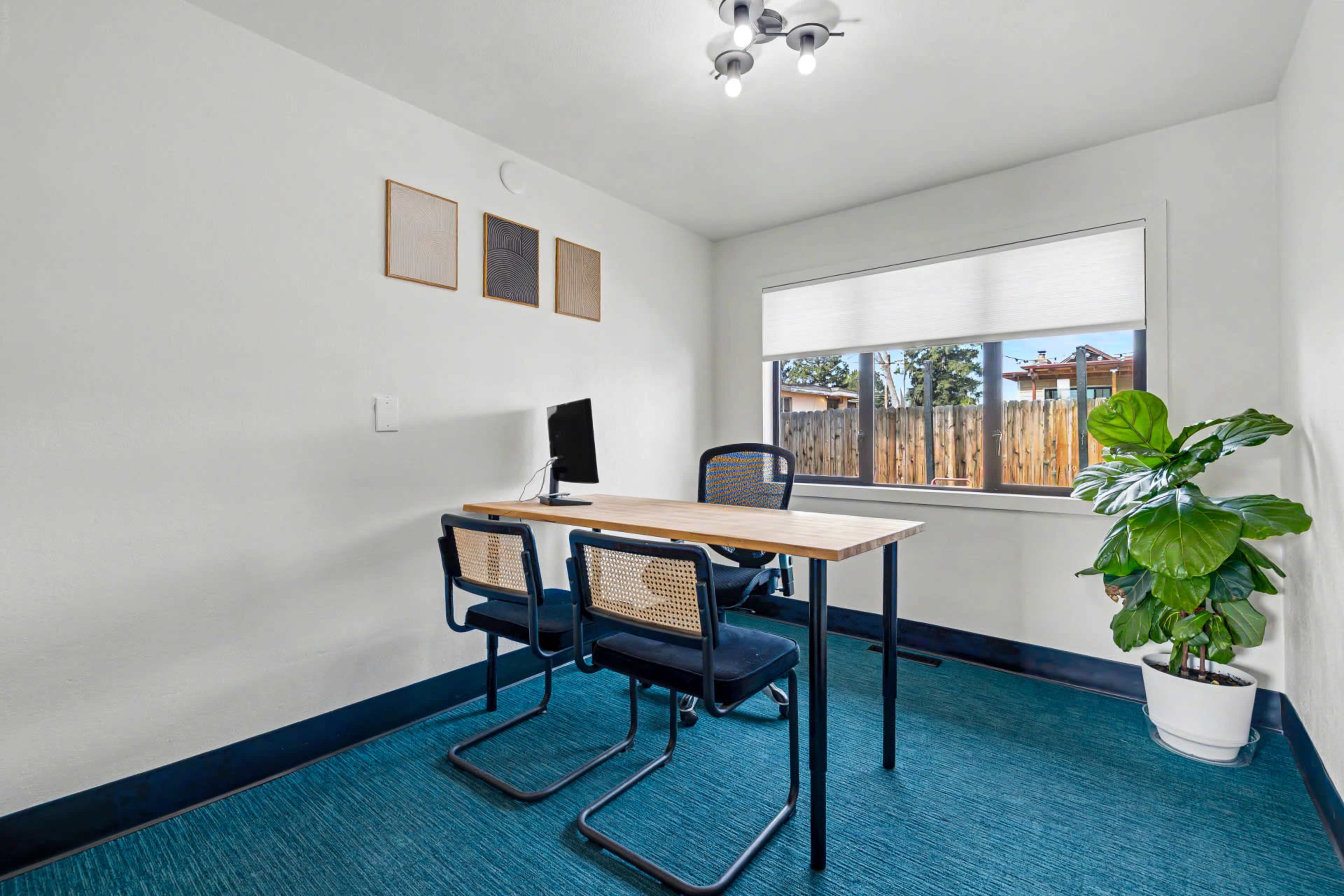 The image shows a small office space featuring a desk with two chairs, a computer monitor, and a potted plant beside a window.