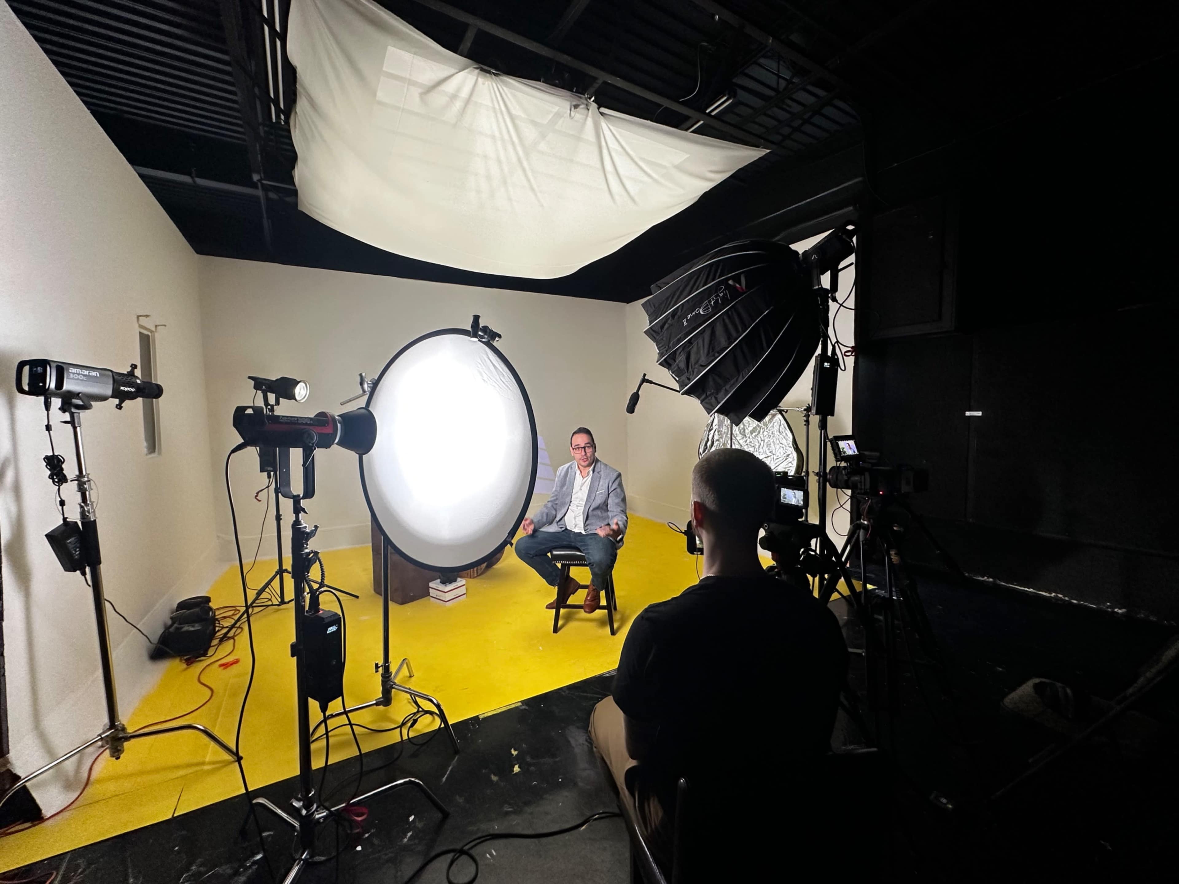 A man sits on a stool in front of bright lights and camera equipment in a studio setting.