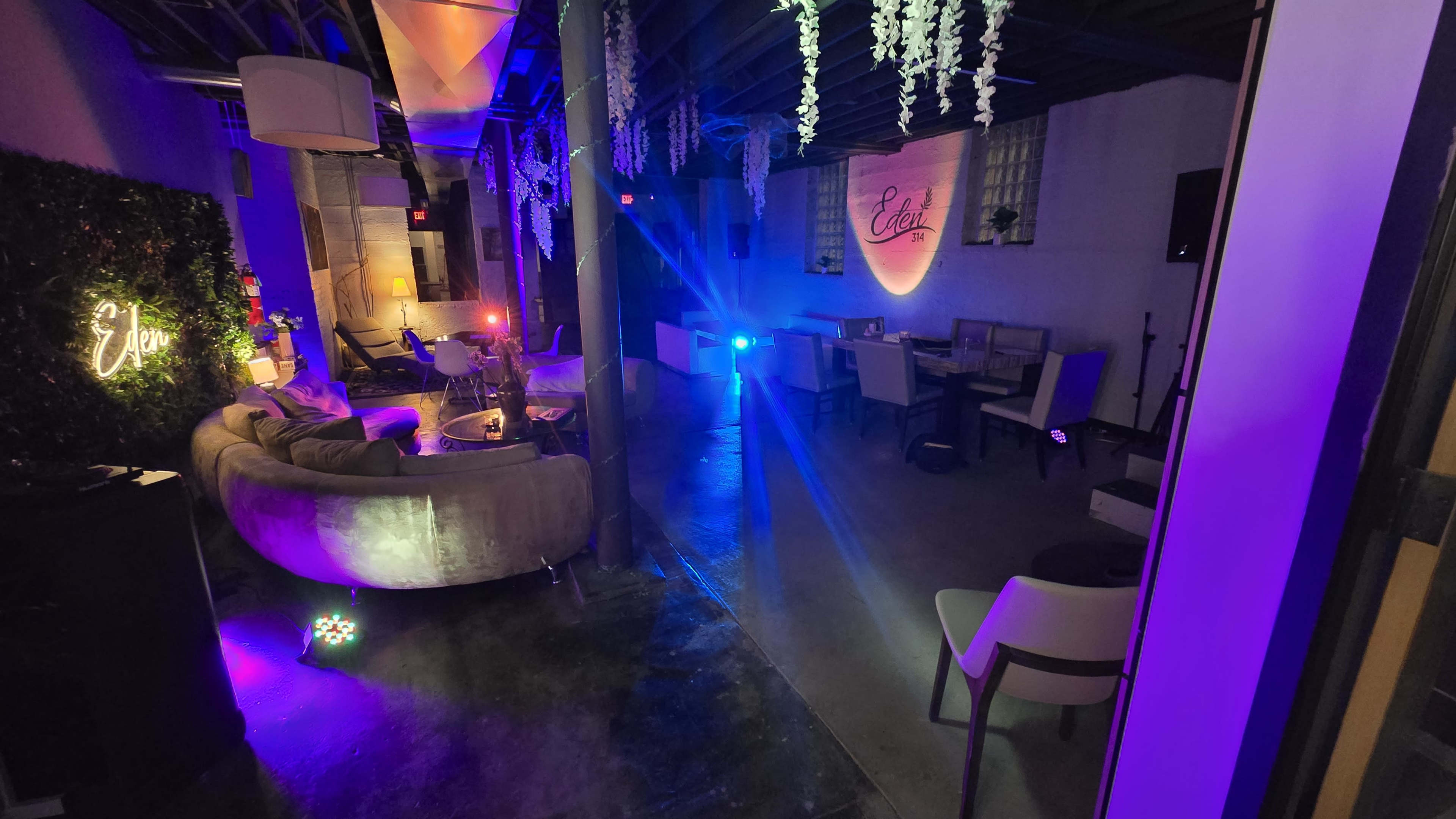 The image shows a dimly lit lounge with colorful lighting, featuring a cozy seating area and decorative elements hanging from the ceiling.