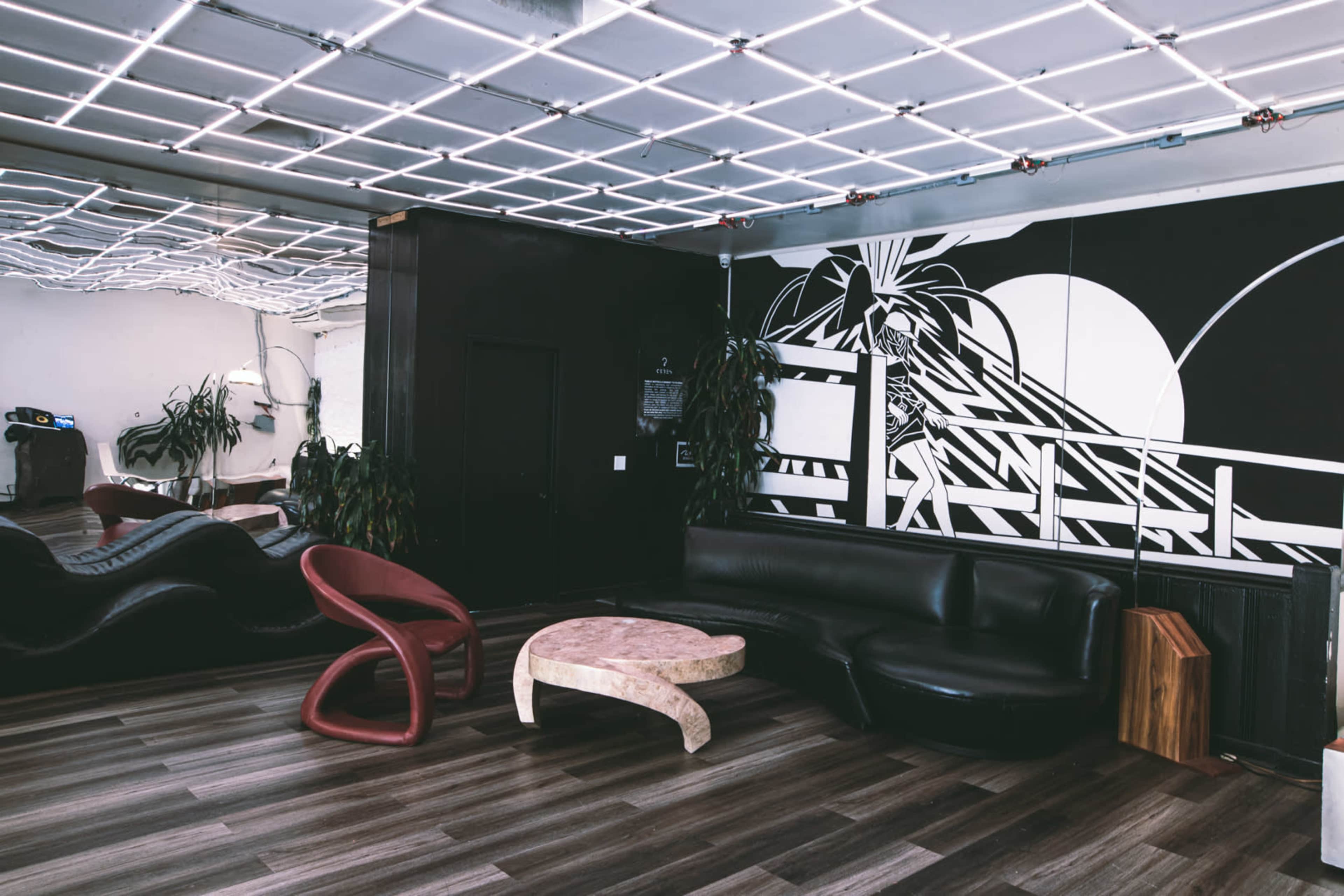 A modern lounge area with a black wall featuring a large graphic design, unique furniture pieces, and a reflective ceiling grid.