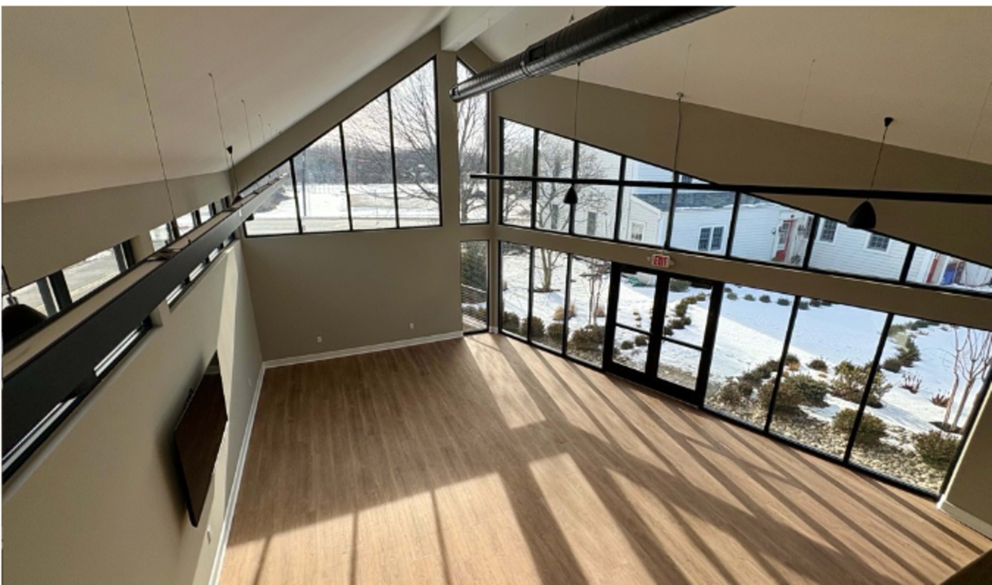 The image shows a spacious, empty room with large windows allowing natural light to enter, featuring a wooden floor and a view of a snowy landscape outside.