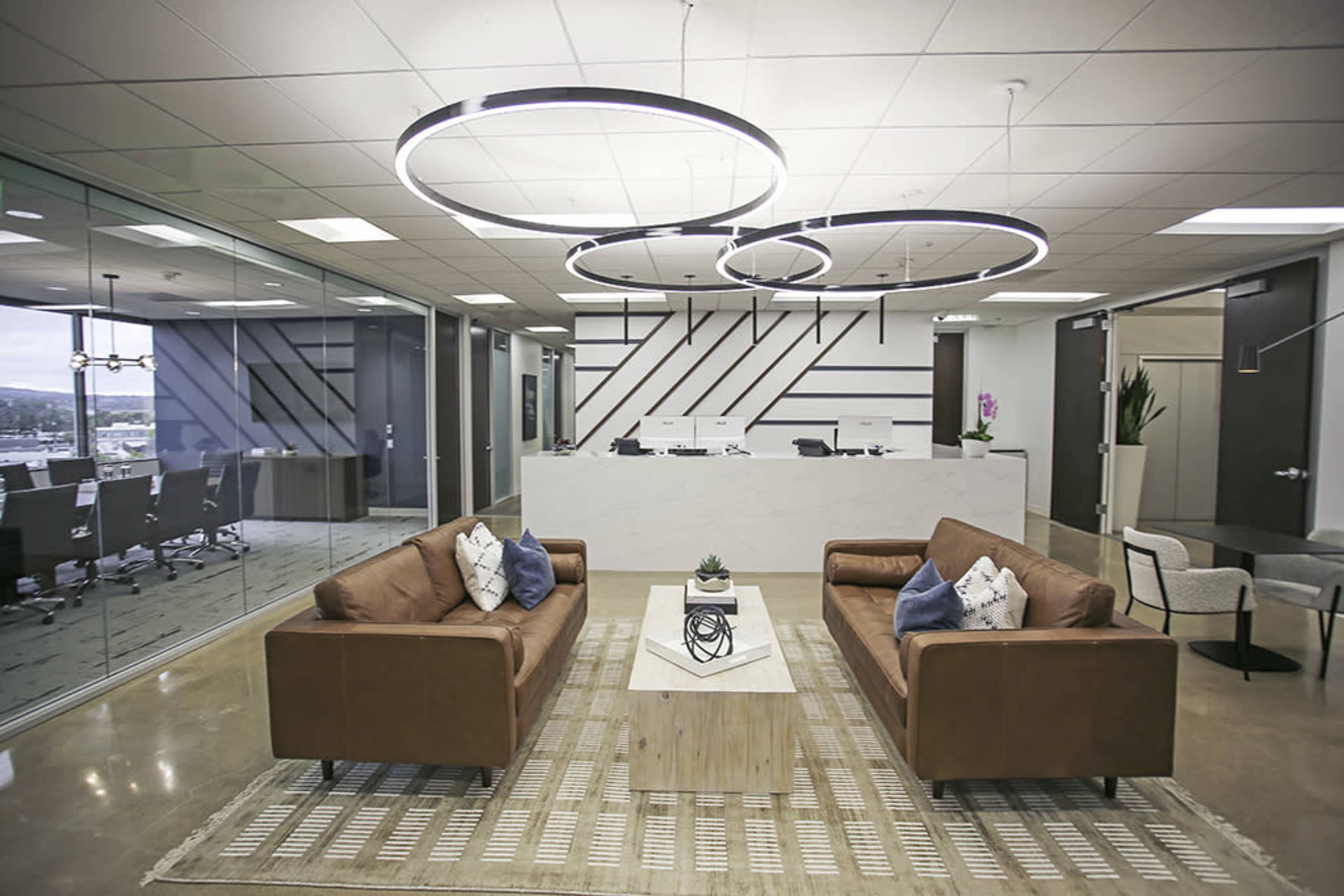 The image shows a modern office reception area with two brown sofas facing a wooden coffee table, surrounded by sleek lighting fixtures and glass walls.