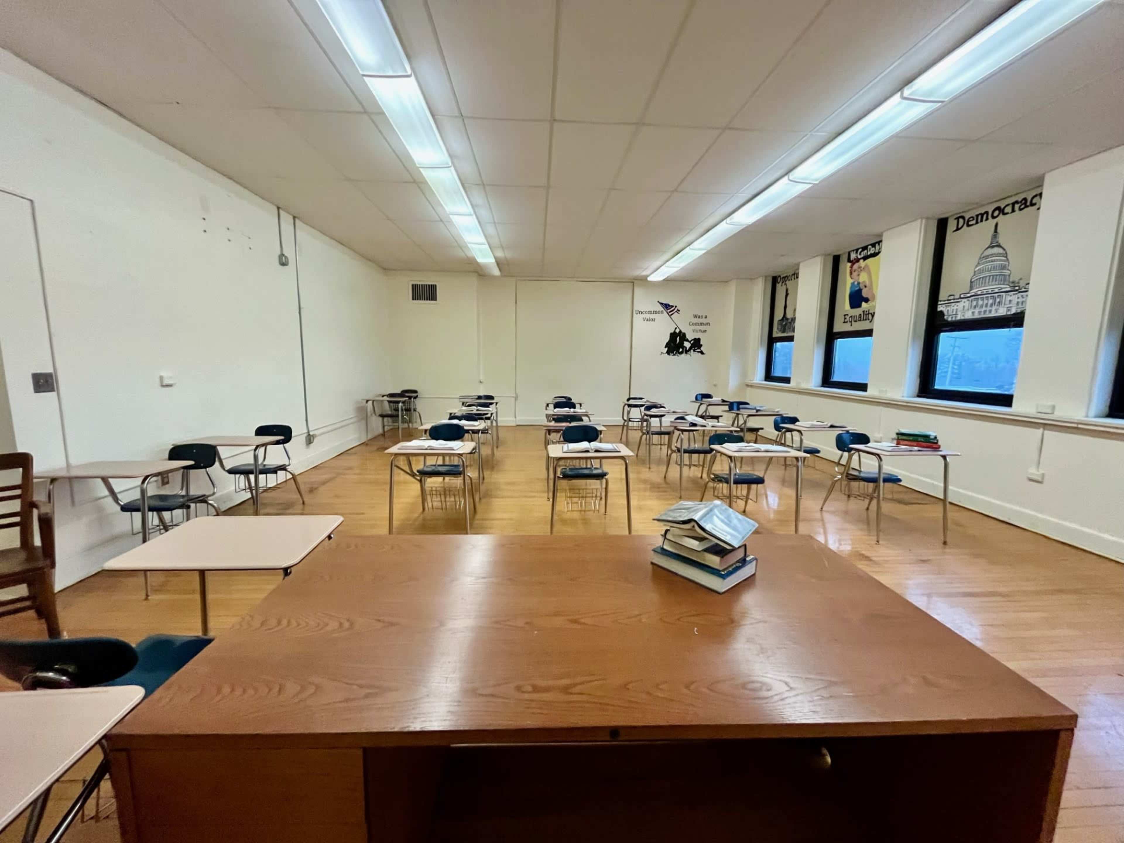 A classroom is set up with rows of desks and chairs, and a large table in the foreground stacked with books.