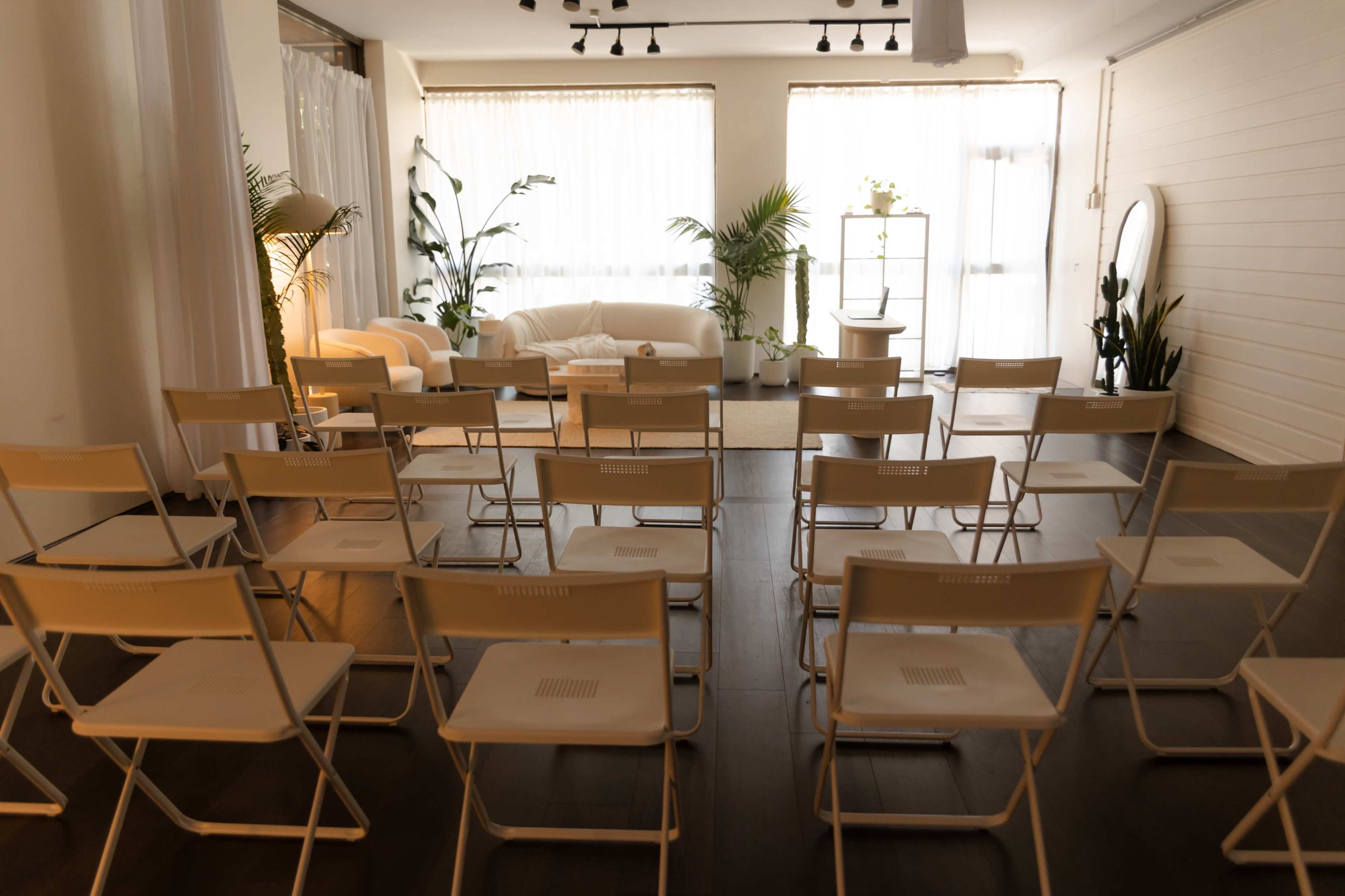 A row of white folding chairs faces a minimalistic setup with a sofa and plants in a well-lit room.