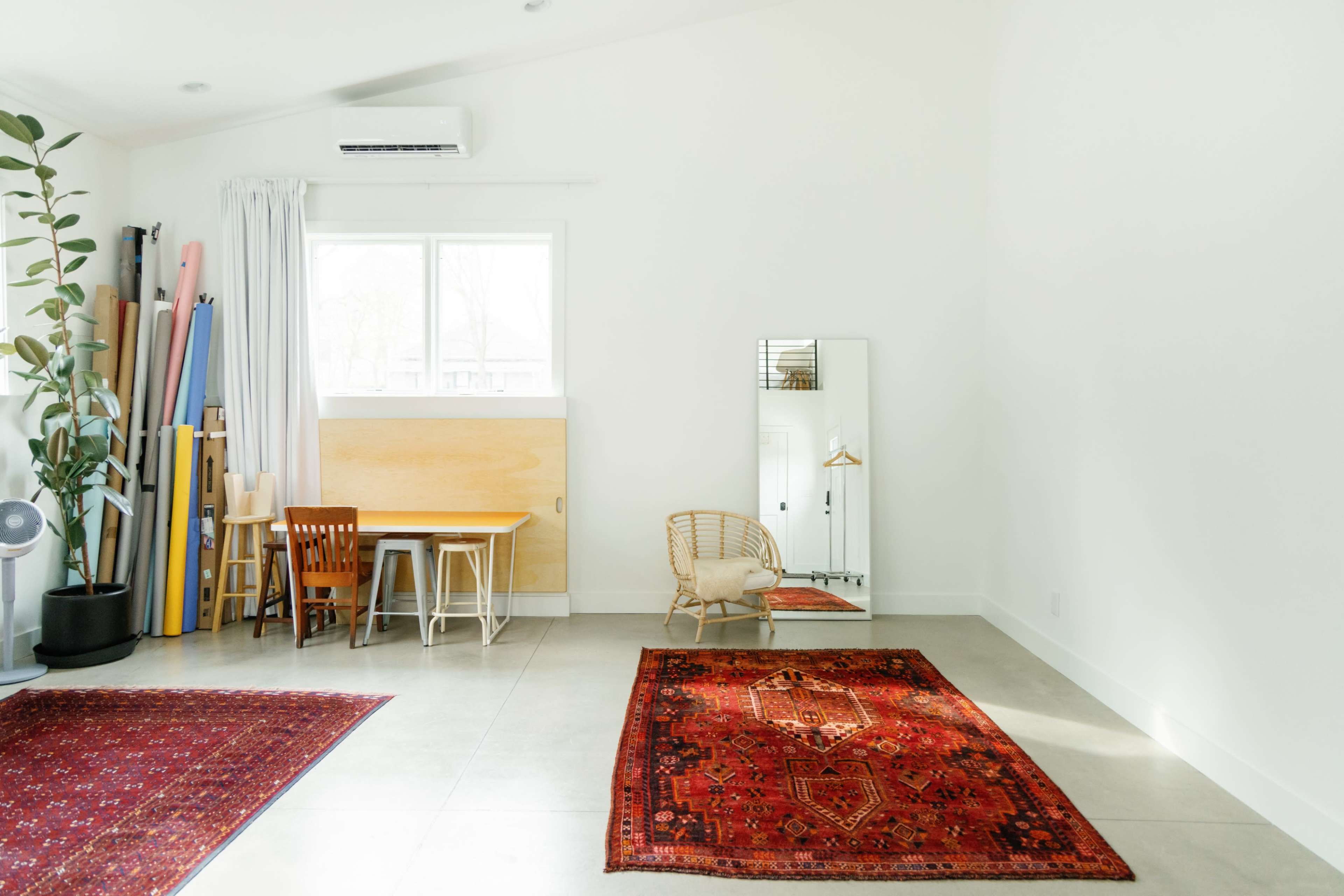 The image shows a bright, minimalist room featuring a dining area with a table and chairs, a large mirror, and two area rugs on a concrete floor.