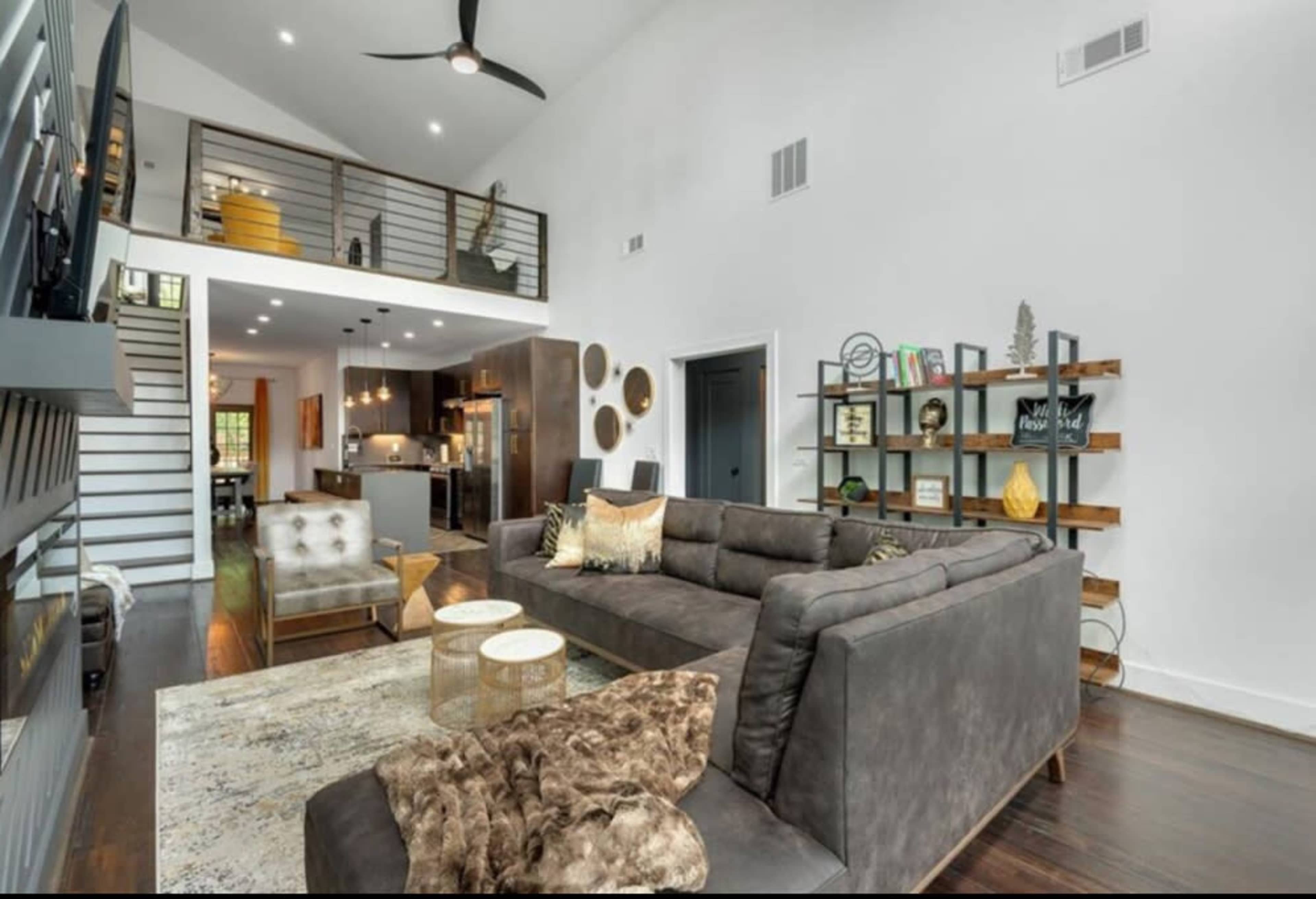The image shows a modern living room with a large, gray sectional sofa, two round coffee tables, and a staircase leading to a lofted area.