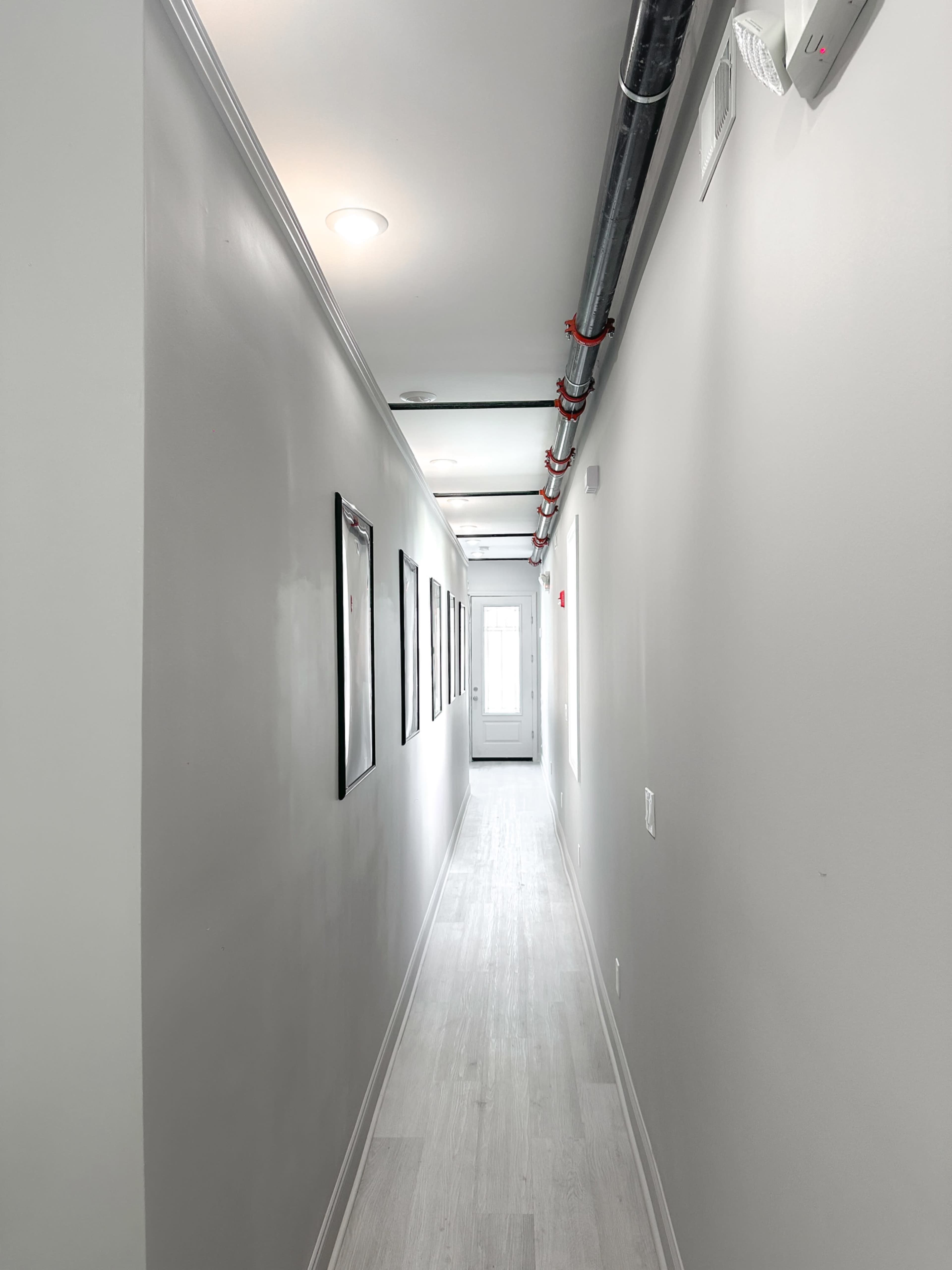 The image shows a narrow hallway with gray walls lined with framed pictures, leading to a door at the far end.