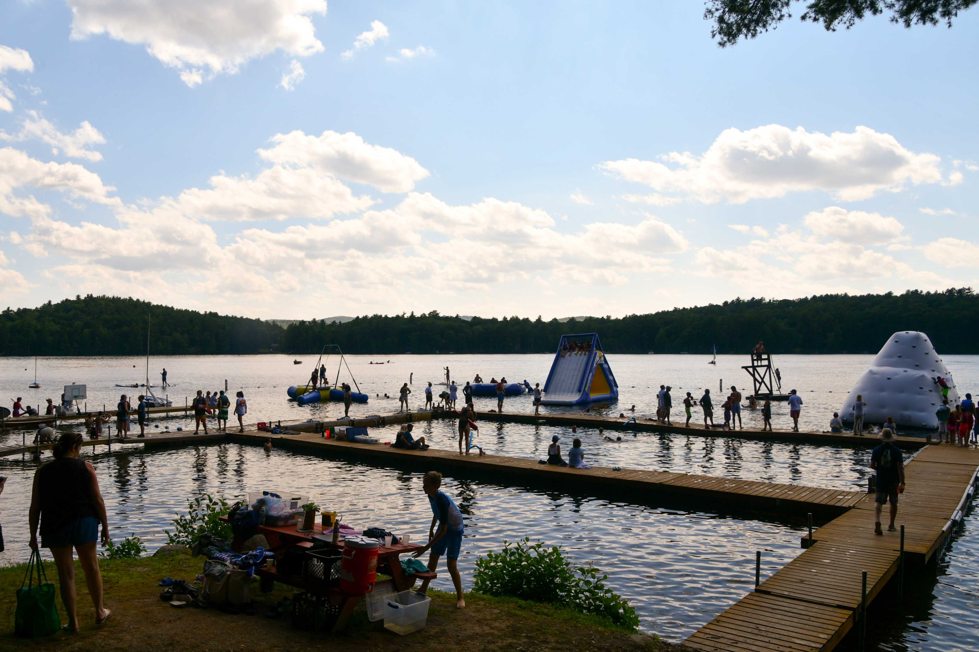 A group of people engages in various water activities along a lakeshore with wooden docks and inflatable attractions.