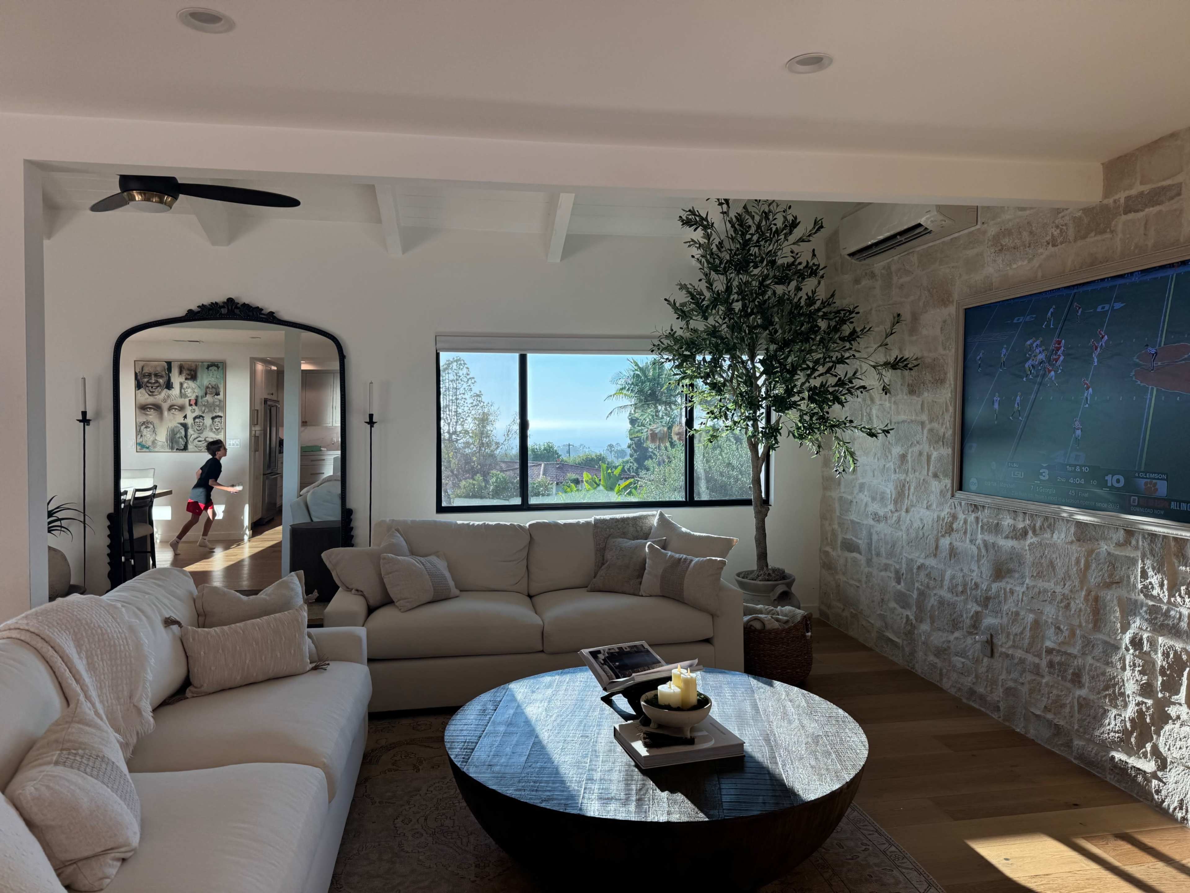 The image shows a bright living room with a large window offering a view of greenery outside, a beige sofa set, a round coffee table, and a television mounted on the wall.