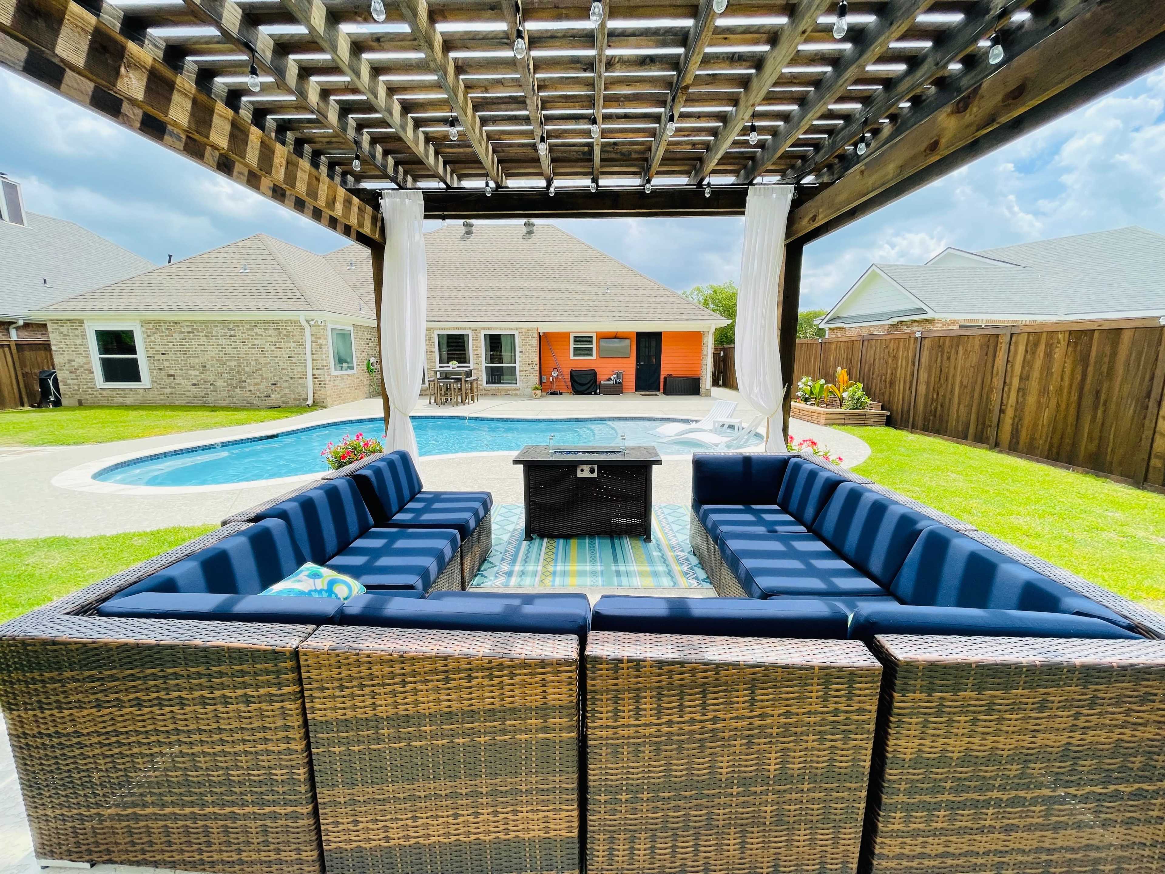 A spacious outdoor patio area features a sectional couch facing a fire pit, with a swimming pool and surrounding homes visible in the background.