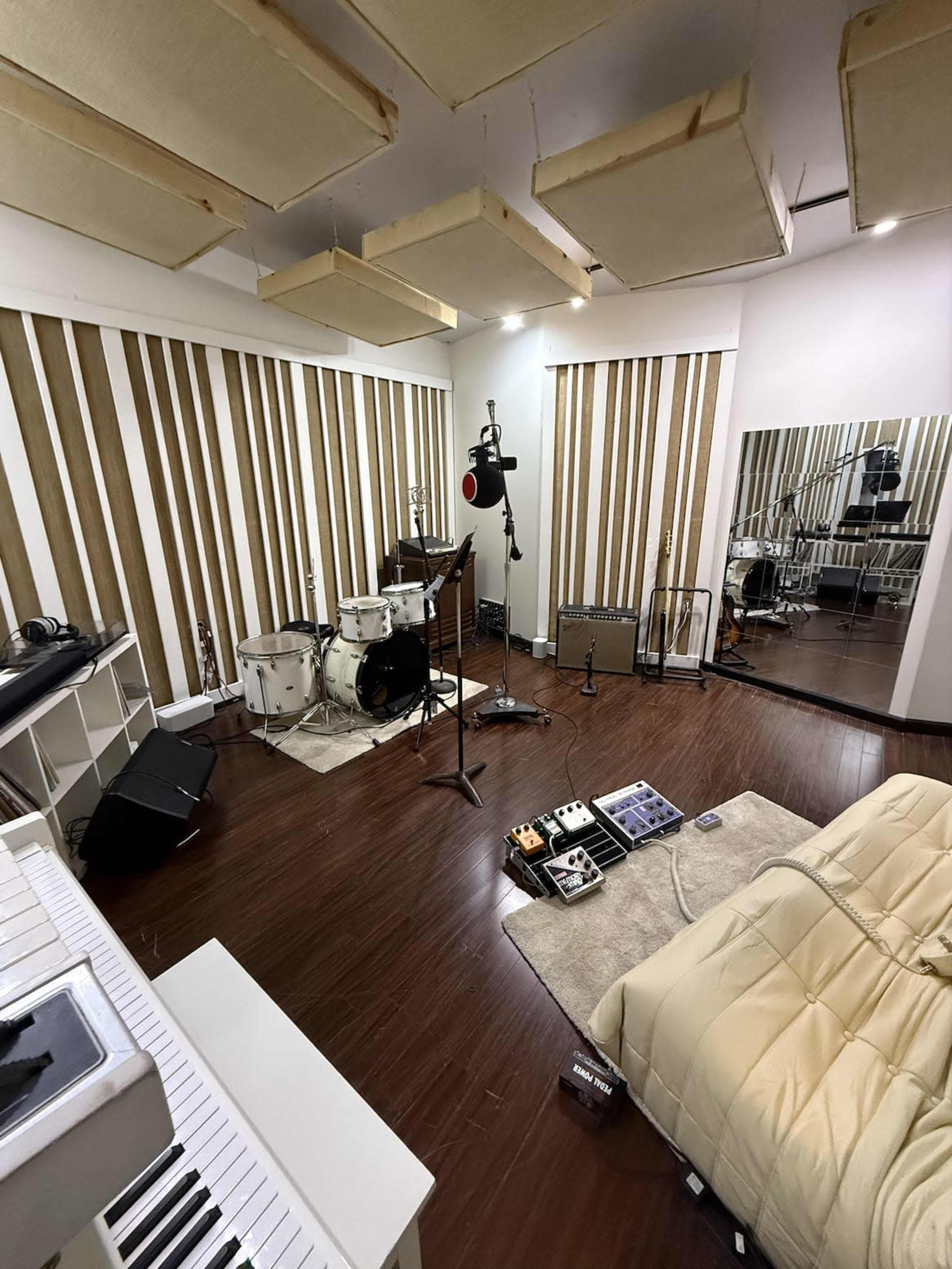 The image shows a music studio with a drum set, various instruments, and a large mirror, all set against striped wooden walls and equipped with soundproofing panels.