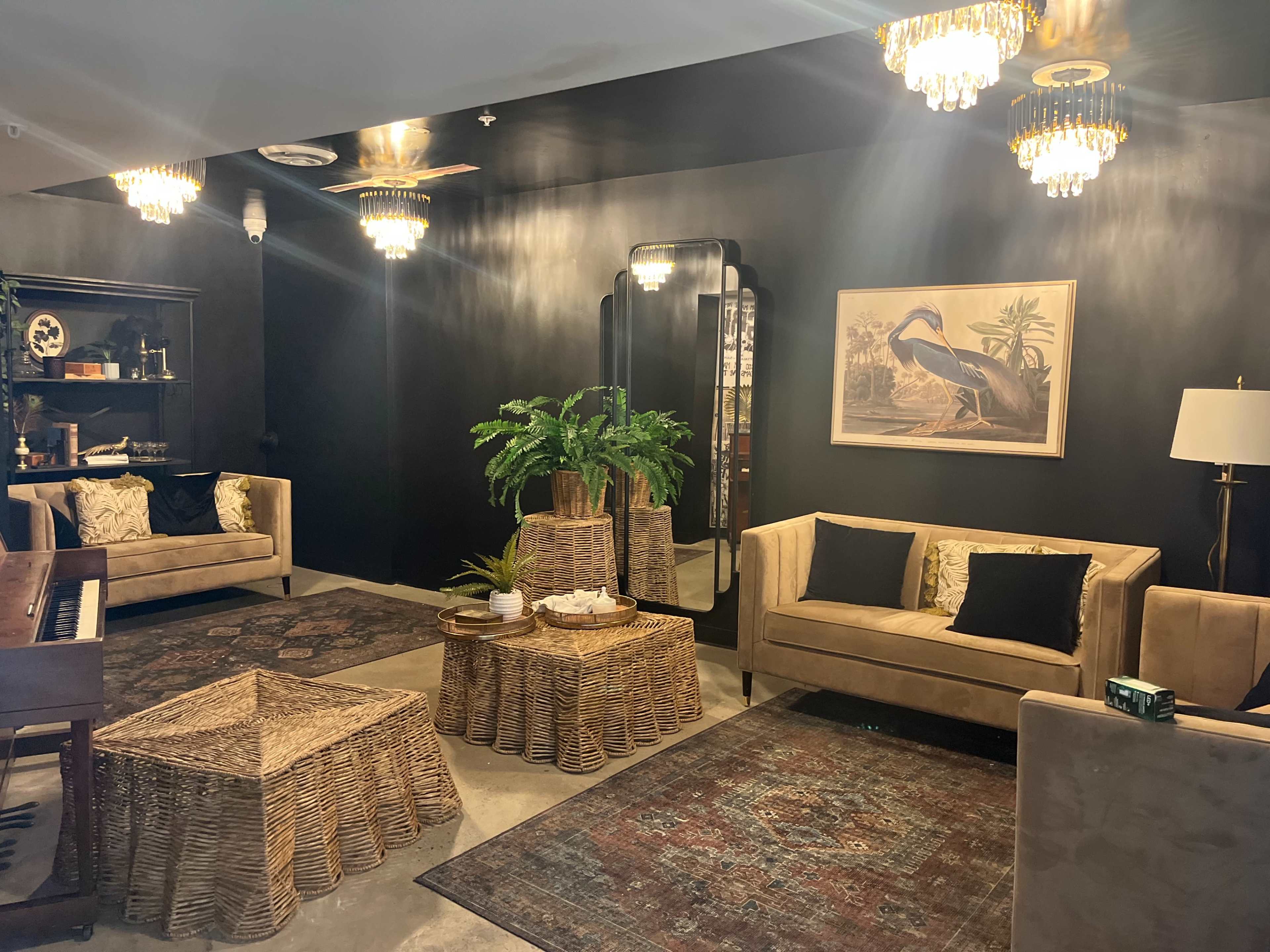 The room features two beige sofas, a piano, a large mirror, and several rattan furniture pieces, all set against dark walls and illuminated by chandelier lighting.