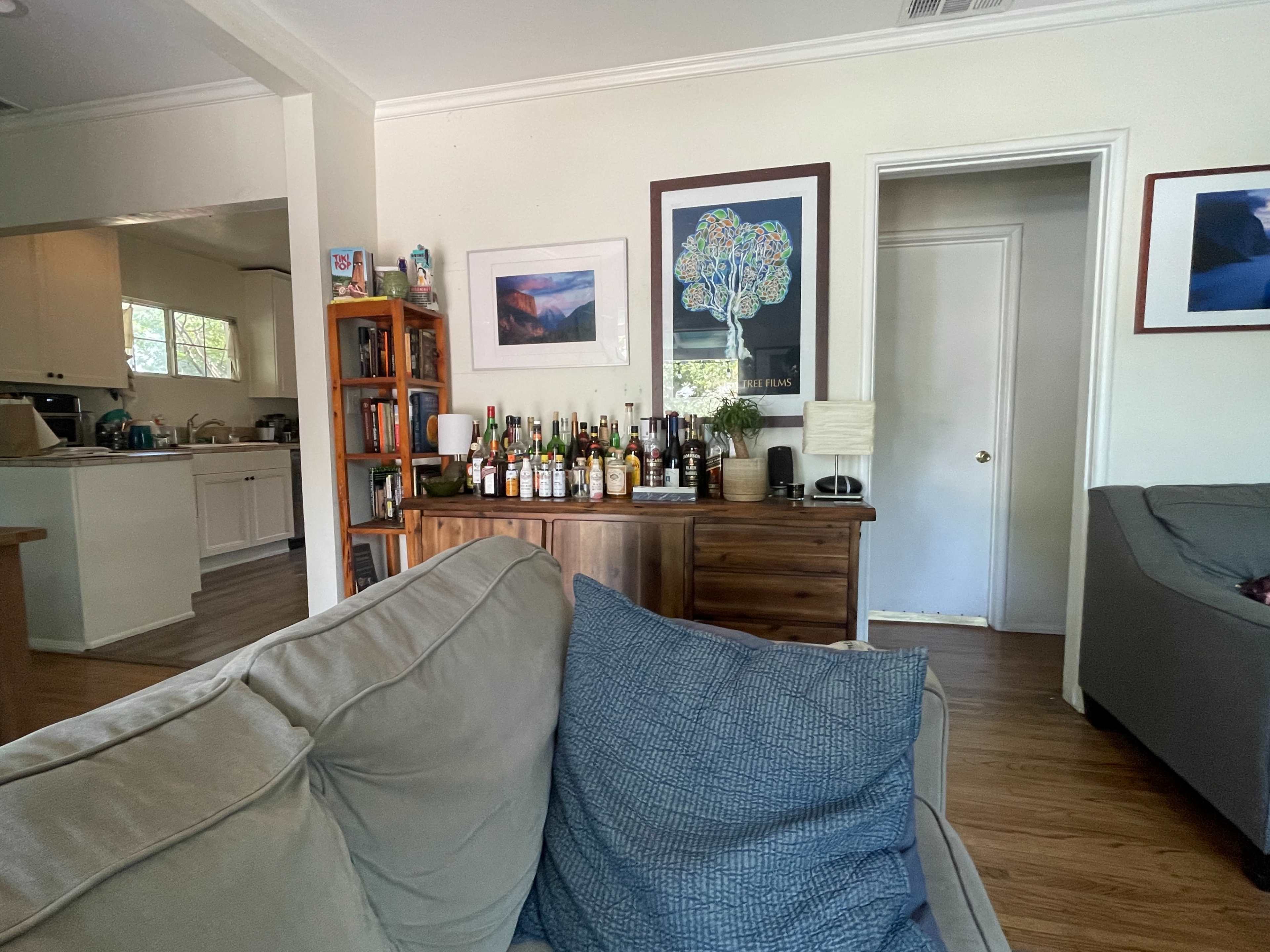 A living room with a couch facing a bar area filled with various bottles, with a doorway leading to another room and kitchen in the background.
