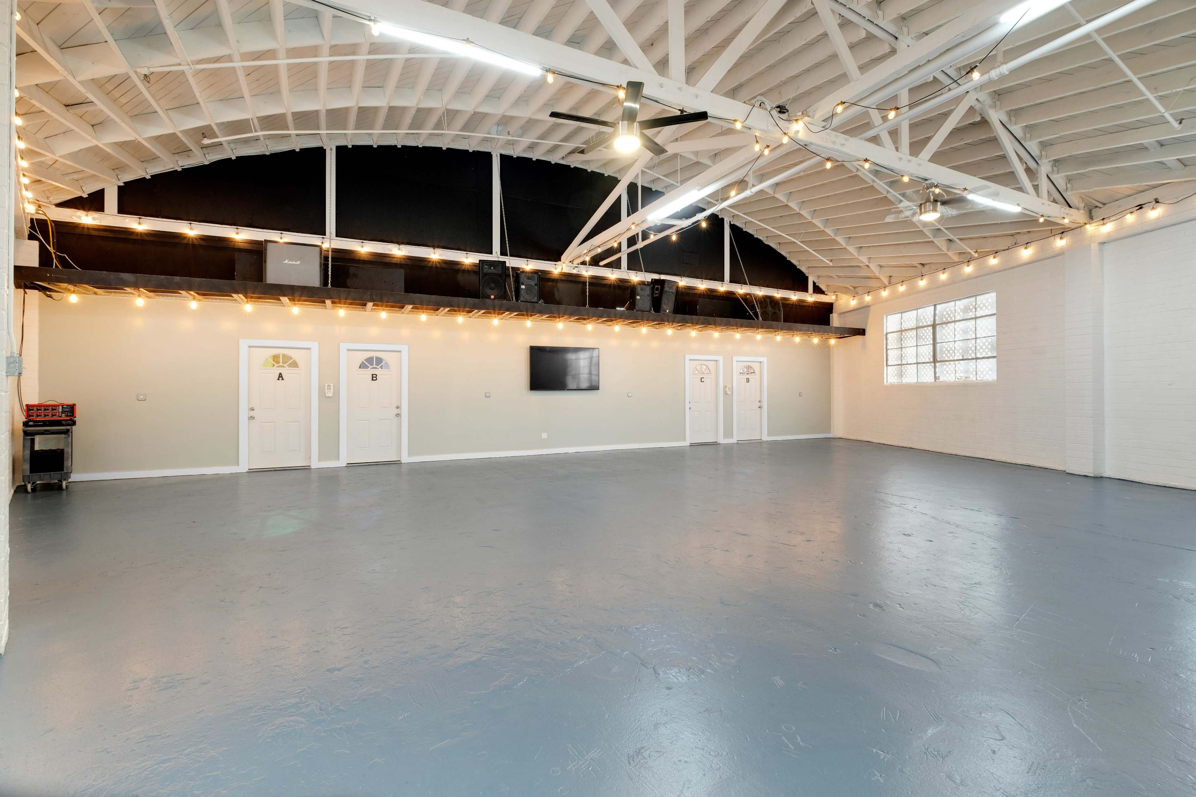 The image shows a spacious room with a high ceiling, featuring multiple doors, a television on the wall, and string lights along the perimeter.