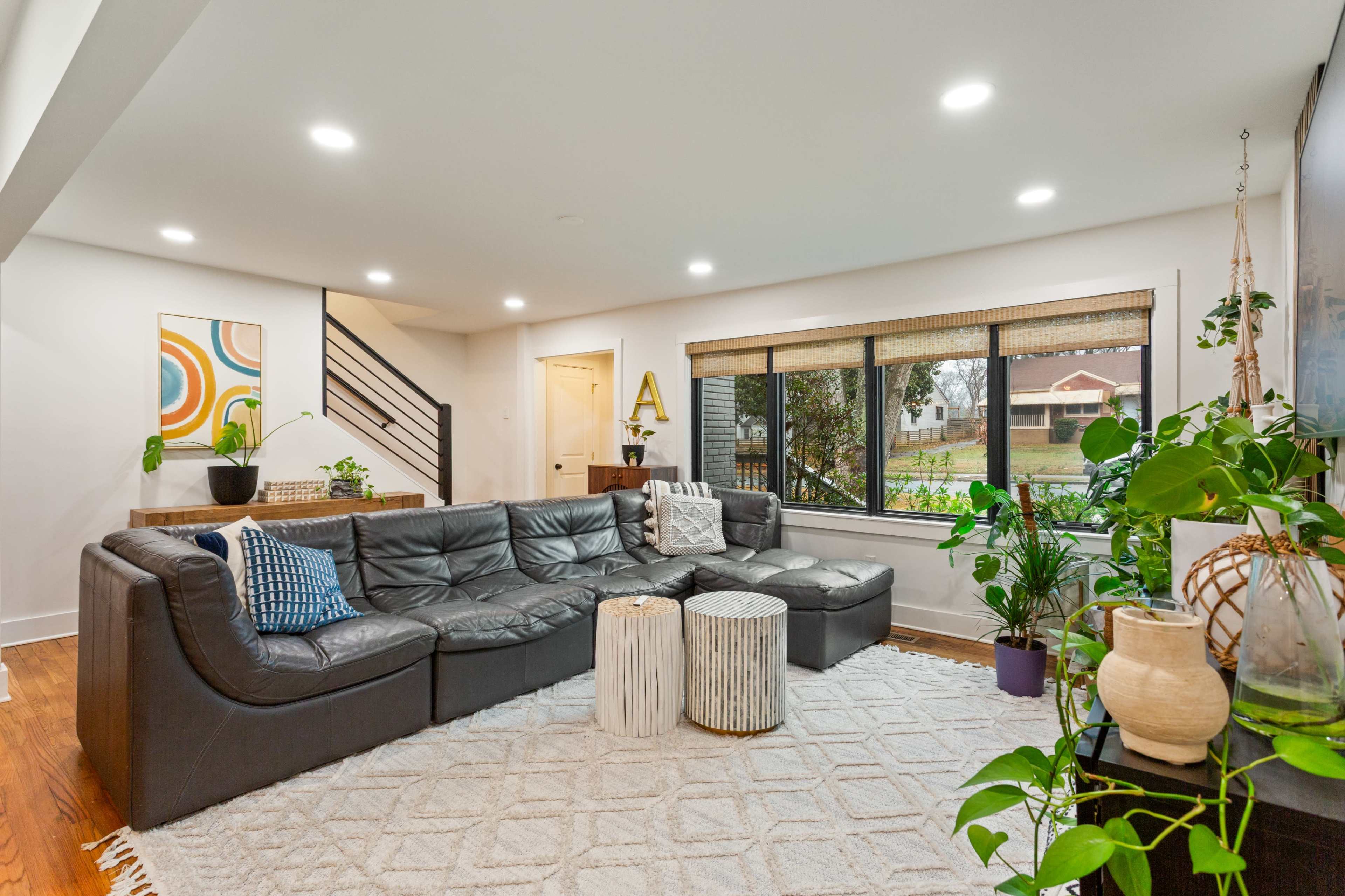 A modern living room features a large sectional sofa, a coffee table, decorative plants, and a window with a view of a garden.