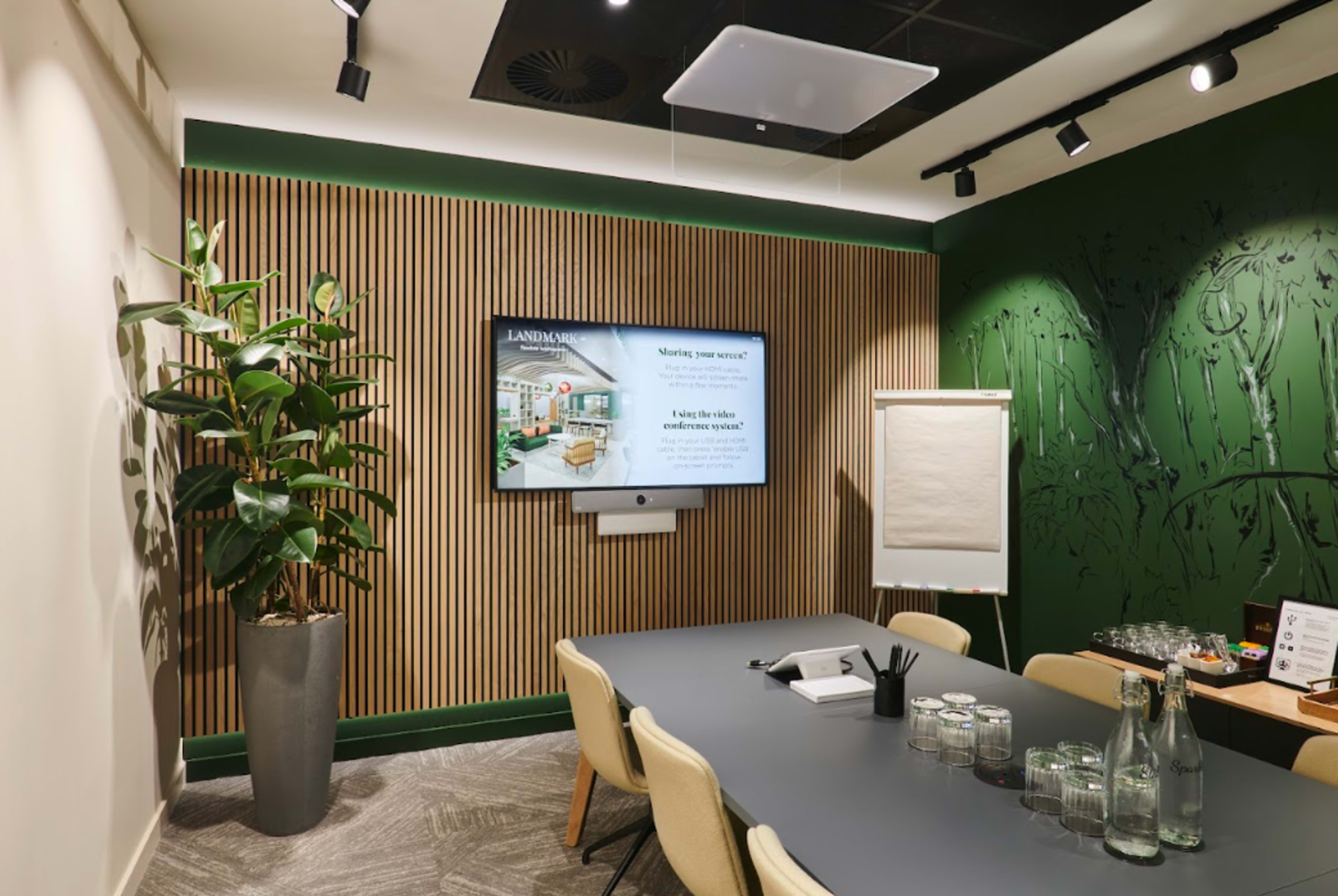 A modern conference room with a large screen, a wooden accent wall, a plant, a table set for a meeting, and a flip chart.