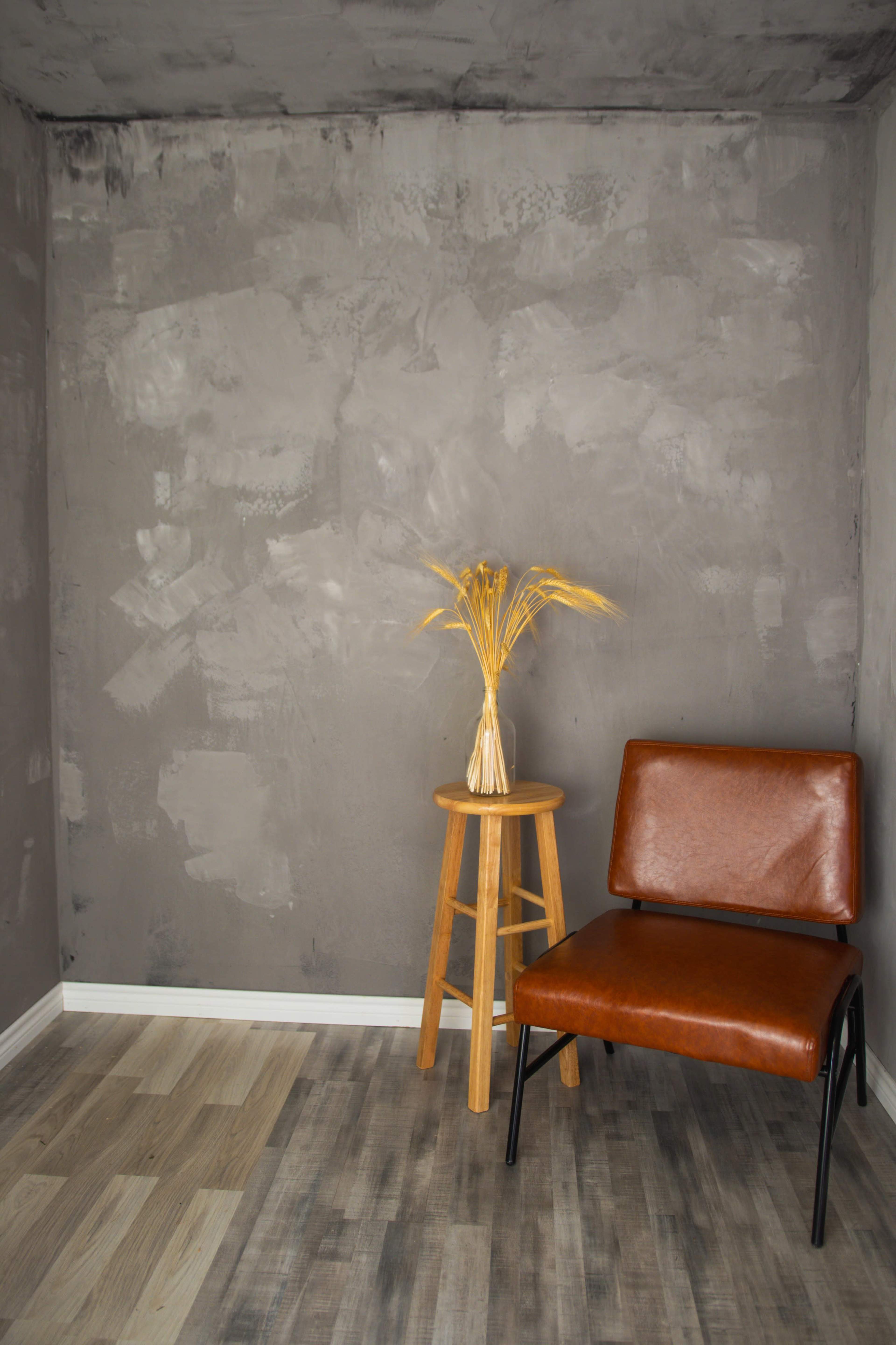 A corner of a room featuring a brown chair next to a wooden stool with a vase of dried wheat arranged on top, against a gray textured wall.